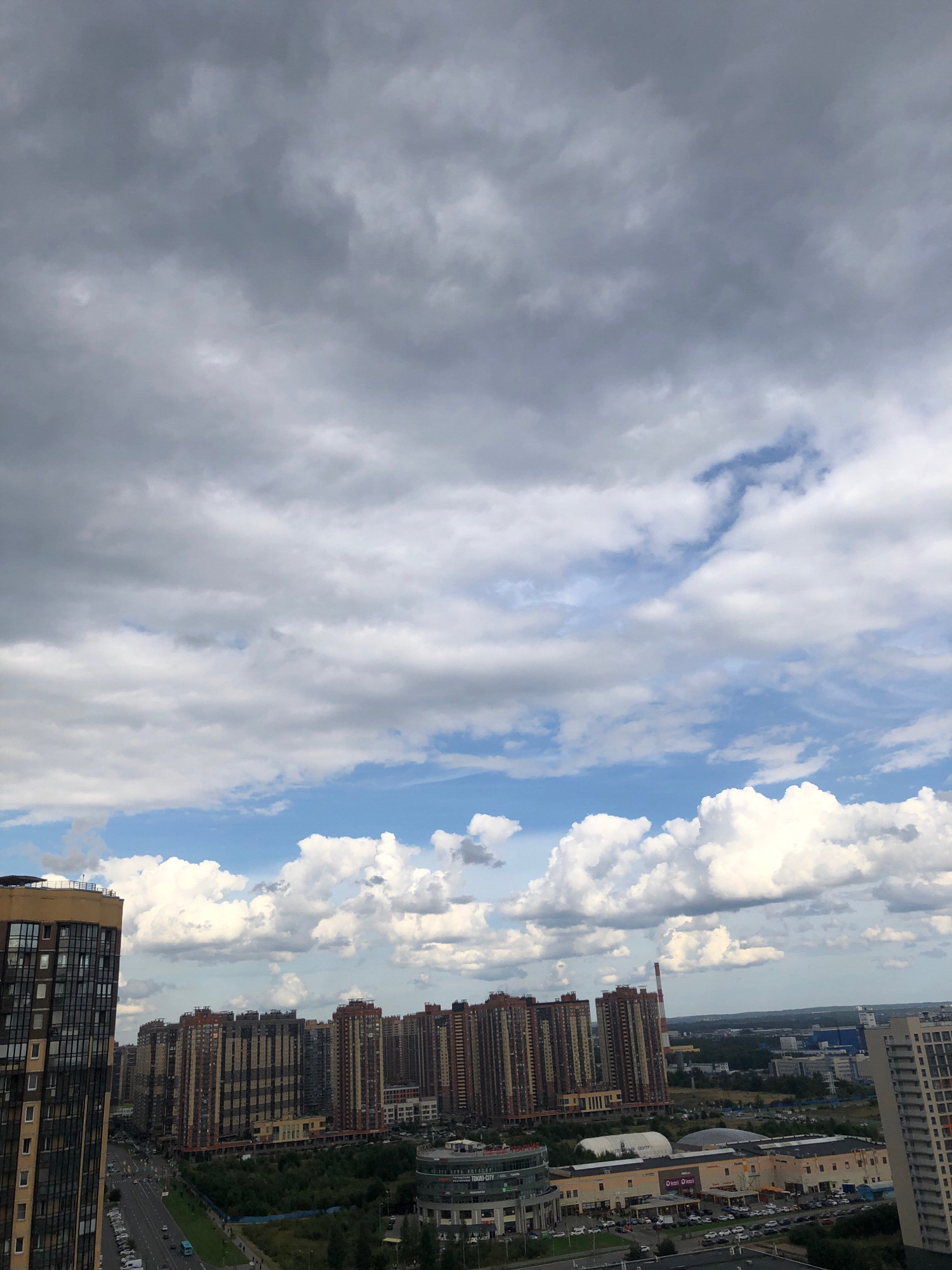 This is a high-angle, eye-level outdoor shot overlooking a modern urban area in an unknown city in Russia. The sky is filled with a mixture of dark and light grey clouds, with patches of blue sky visible. In the foreground, a tall, modern apartment building with a tan and dark facade is on the left. Further back, a row of tall, multi-story apartment buildings with reddish-brown and beige facades dominates the middle ground. These buildings are densely packed, with some showing signs of wear. A wide, tree-lined street with traffic and parked cars runs in front of these buildings. In the background, there are more buildings, including a tall, white industrial smokestack with red stripes. The scene appears to be a residential and commercial district with a mix of high-rise apartments and commercial buildings. The overall impression is one of a bustling, modern city under a cloudy sky.