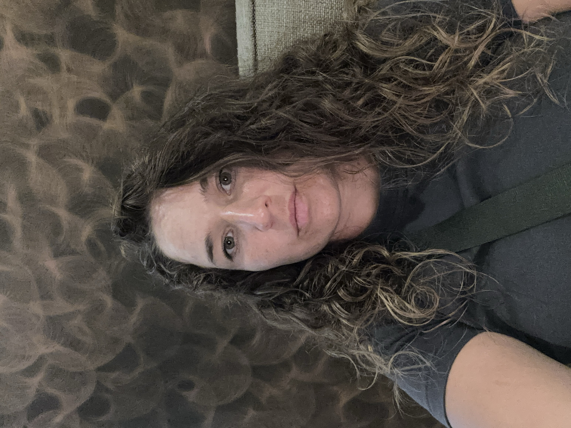A woman with long, curly brown hair takes a selfie indoors. She is wearing a dark grey t-shirt and has a dark green strap across her chest. The background features a textured, patterned wall with circular designs in shades of brown and grey, and a portion of a upholstered chair is visible on the right. The image is a medium shot, focused on the woman's face and upper body. The location is not discernible from the image.