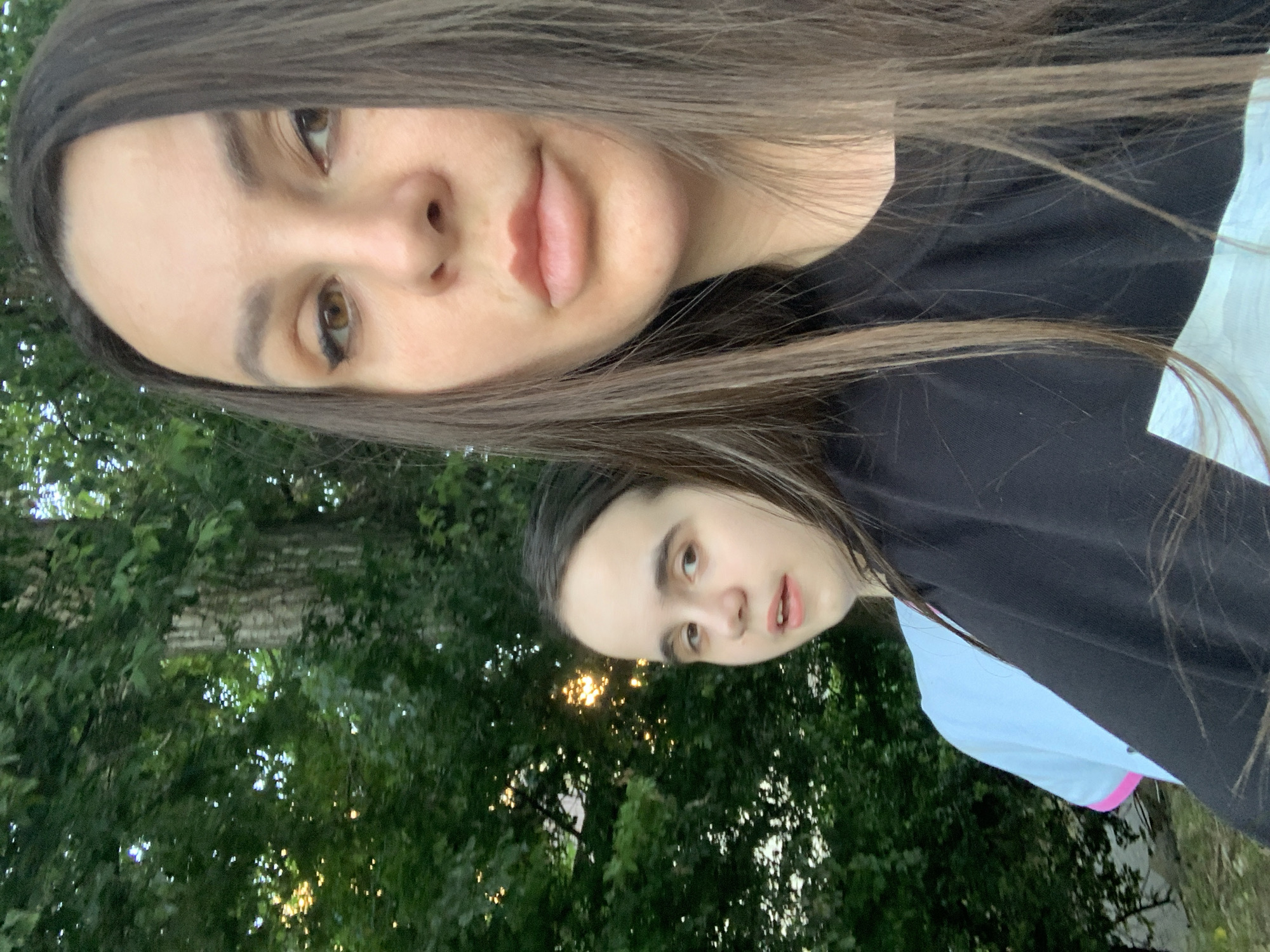 Two individuals are captured in a selfie outdoors, with lush green foliage and trees forming the background. Sunlight filters through the leaves, creating dappled light. The person in the foreground, in focus, is a woman with long, dark brown hair, wearing a black t-shirt. Her gaze is directed towards the camera, and she has subtle makeup on, including eyeliner and lipstick. Behind her, slightly out of focus, is a younger individual with long, dark brown hair, wearing a light blue t-shirt with pink trim. This individual is looking slightly away from the camera with their mouth open. The overall setting suggests a casual outdoor encounter, possibly in a park or natural area within an urban environment.