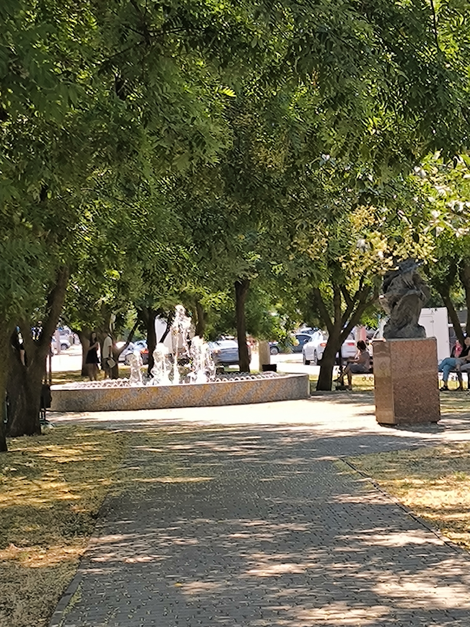The image depicts a park scene in Mykolaiv, Ukraine, on a sunny day. In the foreground, a paved pathway leads into the scene, bordered by grass and dappled with sunlight and shadow from the surrounding trees. To the left, a decorative fountain with water jets is situated within a tiled basin. In the background, several people are visible, some standing near the fountain and others seated on benches. A bronze statue on a granite pedestal stands to the right. Vehicles are present on a road in the distance. The environment is characterized by lush green trees providing shade.