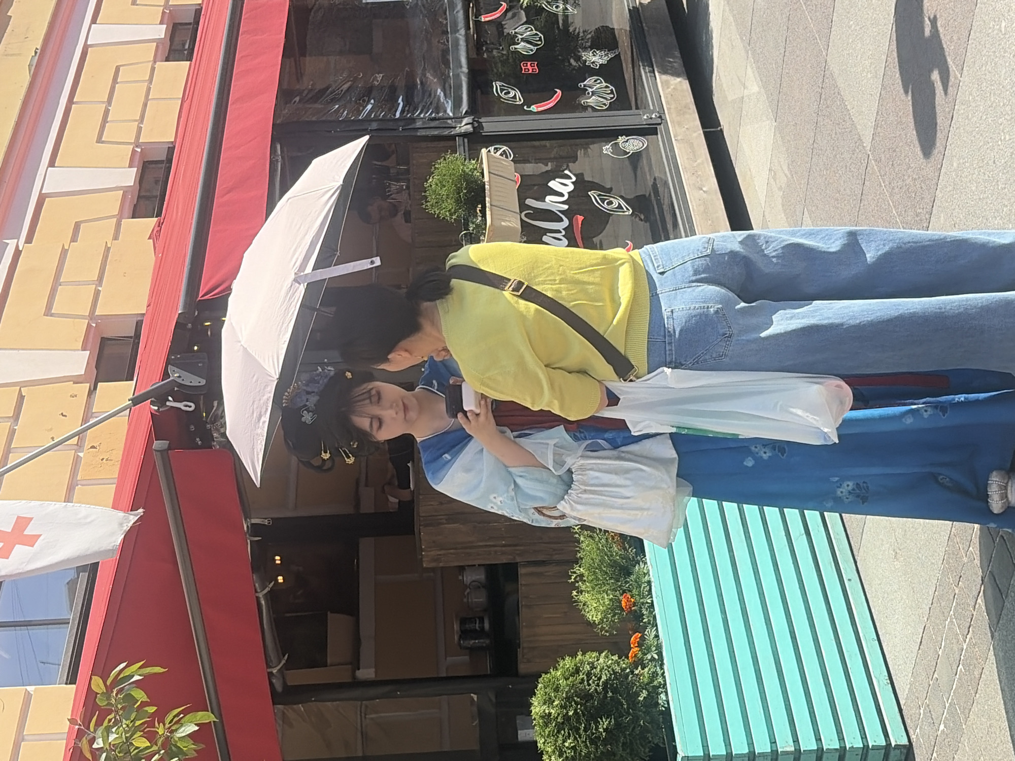 Two women are standing on a city sidewalk in front of a cafe. The woman on the left is wearing a traditional Chinese-style blue dress with a floral pattern, a white bag on her shoulder, and an elaborate hairpiece. The woman on the right is wearing a yellow sweater and wide-leg jeans, and is holding a white umbrella over both of them. They are both looking down, possibly at something one of them is holding. The cafe has a red awning and a glass facade decorated with drawings of food items. A turquoise planter with greenery and flowers is visible to the left of the women. A white flag with a red cross is hanging from a pole above the cafe. The background shows a yellow building with windows. The scene appears to be on a sunny day. This location is likely Saint Petersburg, Russia, given the red cross flag which is commonly associated with medical services or a specific organization in Russia.