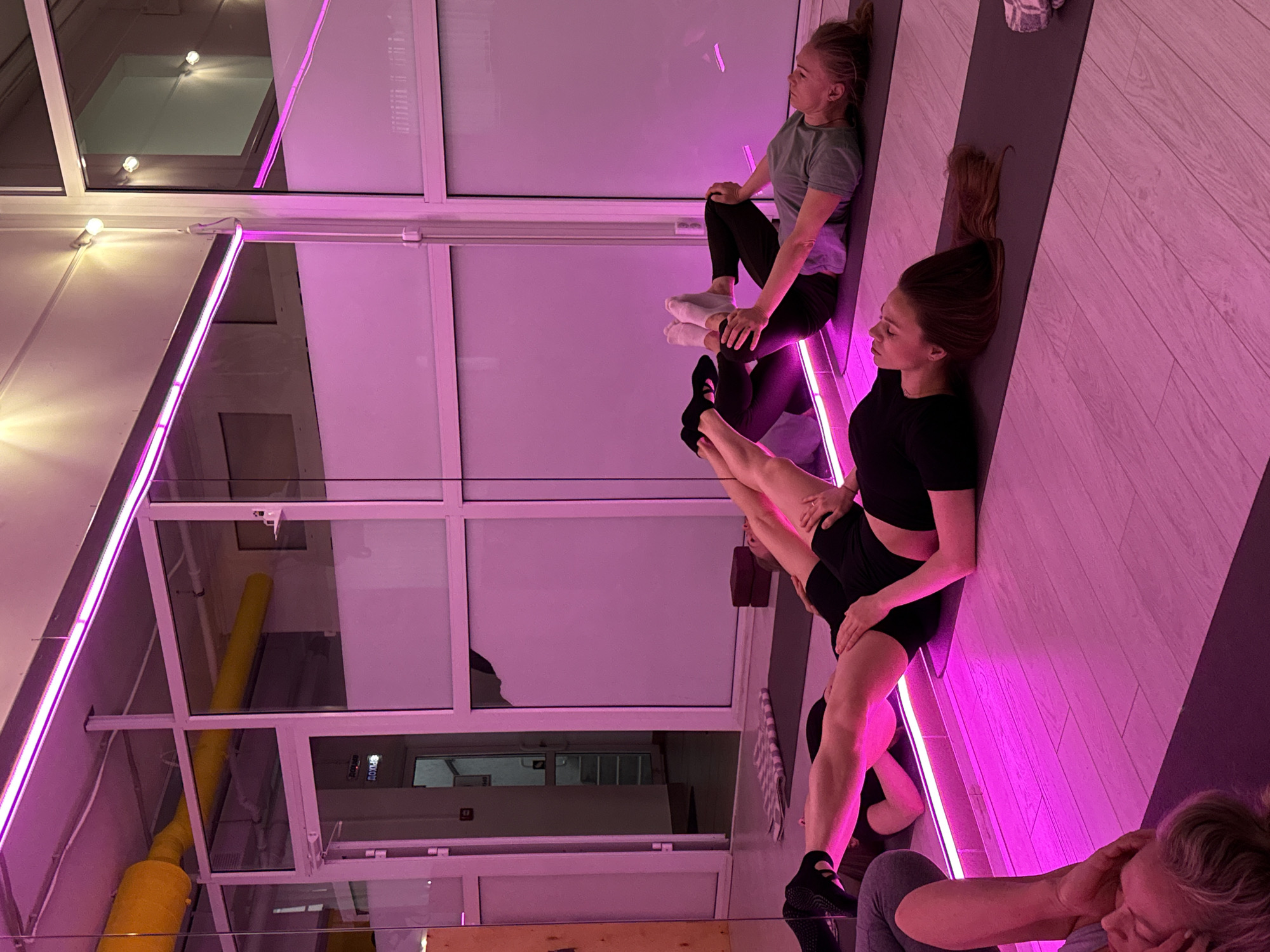 This media file captures an indoor fitness or yoga class, likely in the evening, characterized by strong pink ambient lighting. The perspective is rotated, showing several women engaged in exercise on dark mats laid on a light-colored wooden floor with a herringbone pattern.
The main subjects are three women in athletic wear. One woman, in a grey t-shirt and black leggings, sits with her back against a vertical surface, looking to her right. Her feet are in white socks. Another woman, dressed in a black top and shorts, lies on her side or back on a mat, her legs bent and raised in a pose, wearing black athletic shoes. She is looking to her left. A third woman, partially visible in the bottom right, wears grey pants and has a hand near her head.
The setting appears to be a modern studio or gym. Prominent features include large white-framed windows or glass panels that offer reflections of the interior, including warm white ceiling lights. A key visual element is the vibrant pink LED strip lighting running vertically along a wall or column, casting a strong magenta glow throughout the scene. Through the glass, a bright yellow cylindrical pipe or ventilation duct is visible.
The overall atmosphere is one of focused activity within a uniquely lit environment. Through one of the glass panels, faint Cyrillic text, possibly "ВЫХОД" (VYKHOD, meaning "EXIT"), can be discerned, providing a subtle location cue from Chaikovsky, Russia.