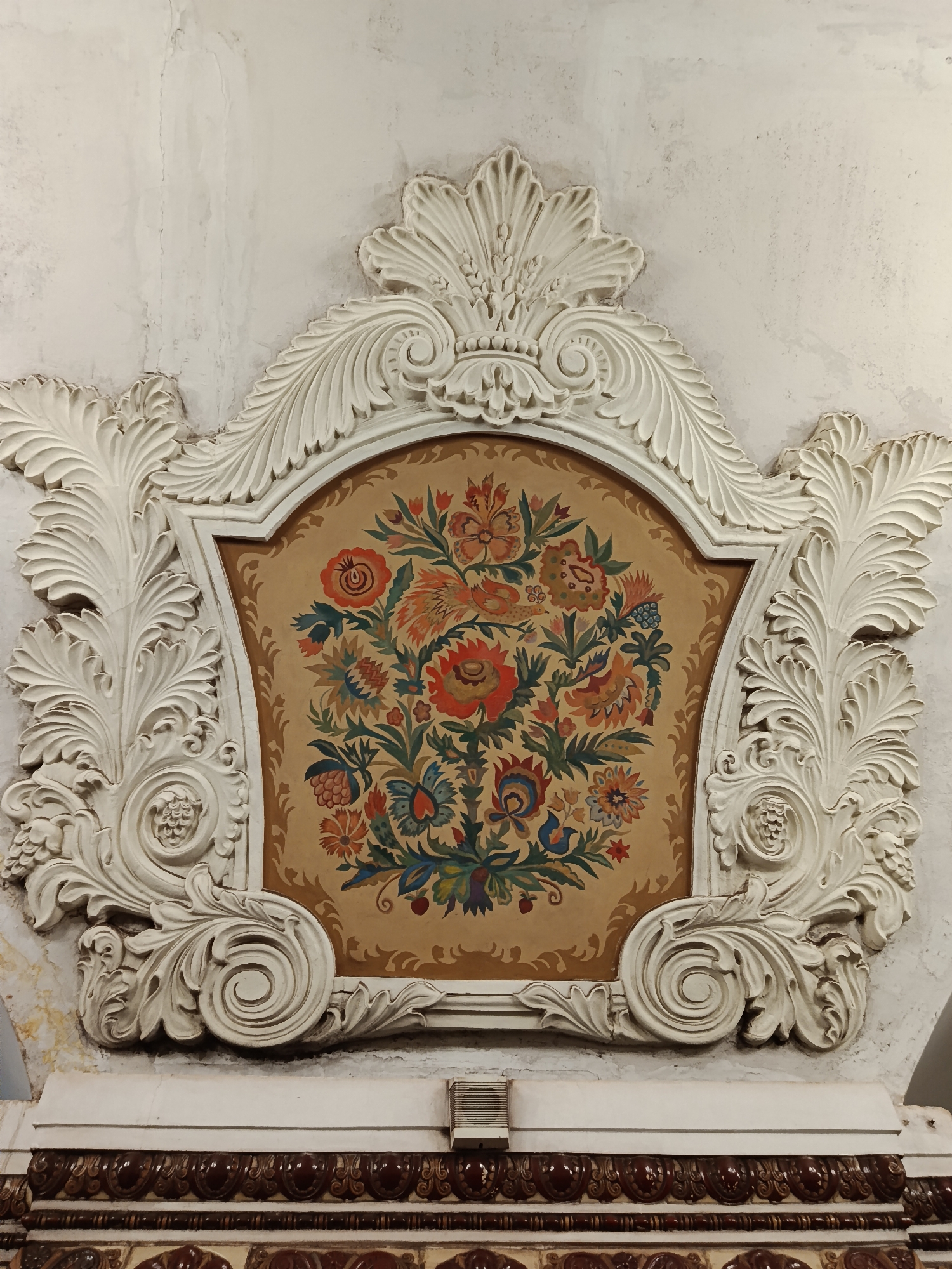 A floral decorative panel is seen on a white wall, likely indoors. The panel features a central tan background with a stylized floral arrangement in various colors, including red, orange, green, and blue. The painted floral artwork is surrounded by an ornate, white, decorative frame with swirling and leaf-like designs. The wall itself appears textured and aged, with evidence of repairs or repainting. Below the panel, there is an additional decorative strip with a brown and white pattern. The image is a detail of a station of the Moscow metro, specifically the Baumanskaya station, in Russia.