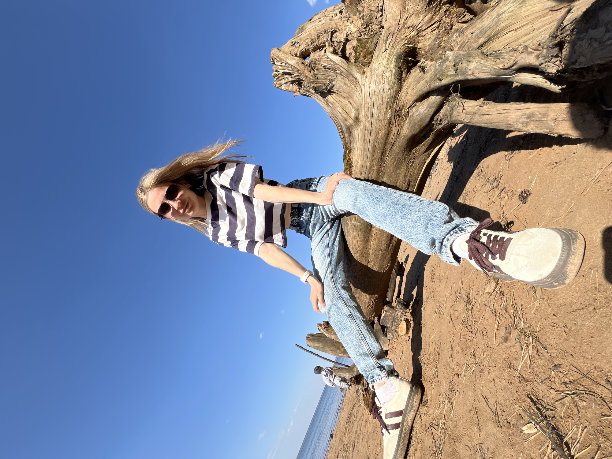 A woman is sitting on a tree stump on a sandy beach. She is wearing a striped top, jeans, and sneakers. The sky is blue and clear. In the background, there is a body of water. There are two people on the beach behind the woman. The location is in the city of Chaikovsky, Russia.