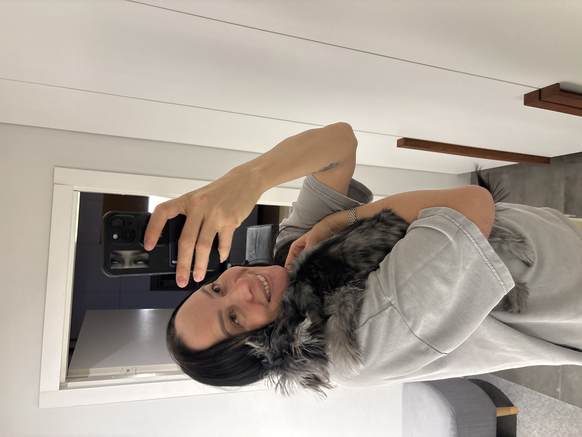 This image depicts an indoor scene in Chaikovsky, Russia. An adult female, visible from the chest up, with dark hair, is smiling and looking towards the camera. She is holding a dark grey, fluffy, feathered animal, likely a Silkie chicken, against her left shoulder and chest. The woman is wearing a light grey, short-sleeved t-shirt, a metallic bracelet on her left wrist, and has a faint tattoo on her right forearm. She is taking a mirror selfie using a black-cased smartphone held in her right hand. The background consists of white walls. To the left, a white door frame opens into a darker room. To the right, a white wardrobe or cabinet with two vertical wooden handles is visible. A grey upholstered stool with light wooden legs is partially visible in the lower-left foreground. The floor appears to be light grey.