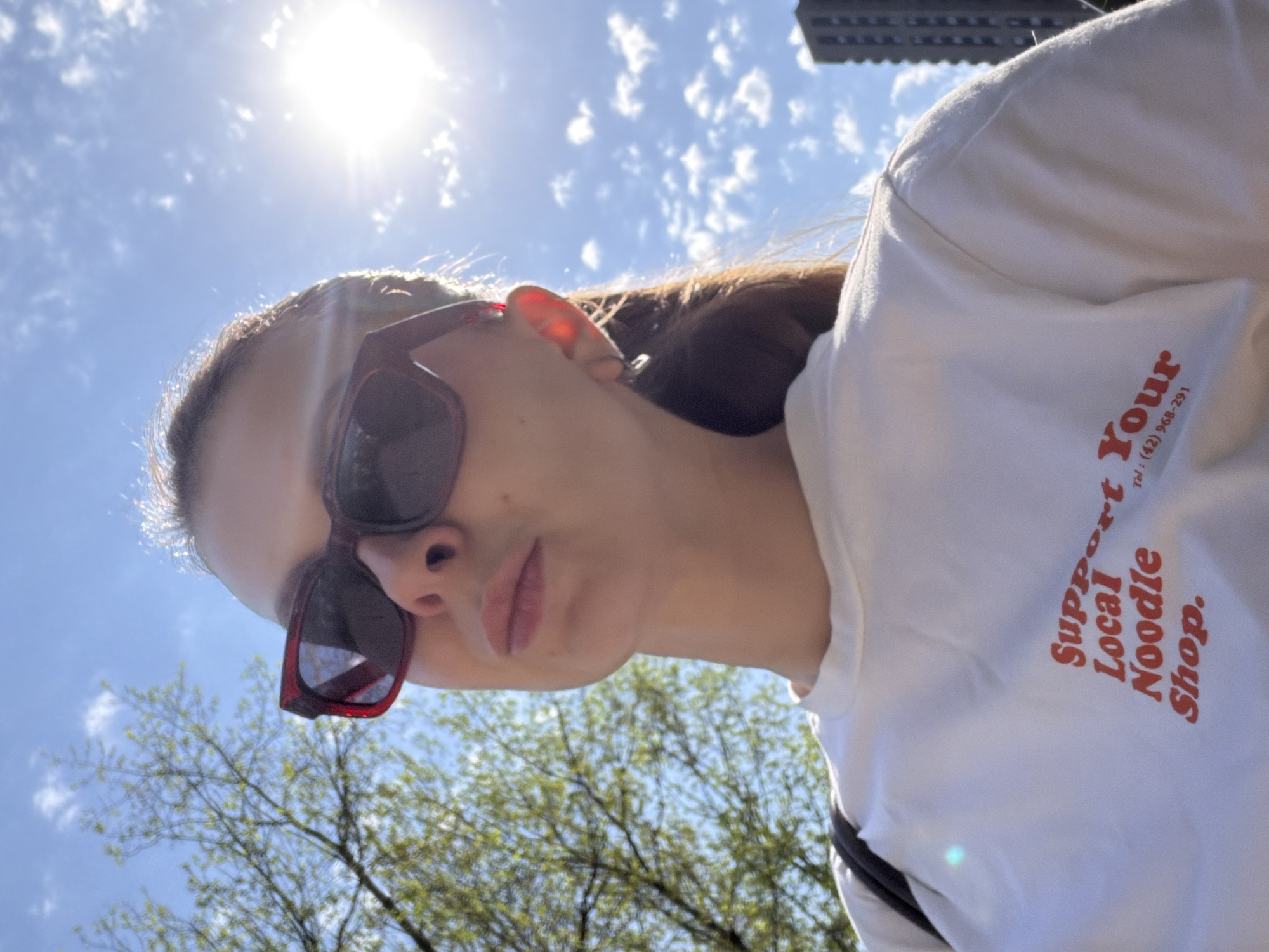 A selfie of a woman wearing sunglasses and a white t-shirt with red text that reads "Support Your Local Noodle Shop. Tel: (42) 968-291" is taken from a low angle on a bright, sunny day. The sun is visible in the upper left corner of the frame, surrounded by a blue sky with scattered clouds. Part of a building is visible in the upper right of the image and branches of trees in the lower left. It's unlikely the photo was taken in Khimki, Russia, given the phone number provided on the shirt is a US phone number.