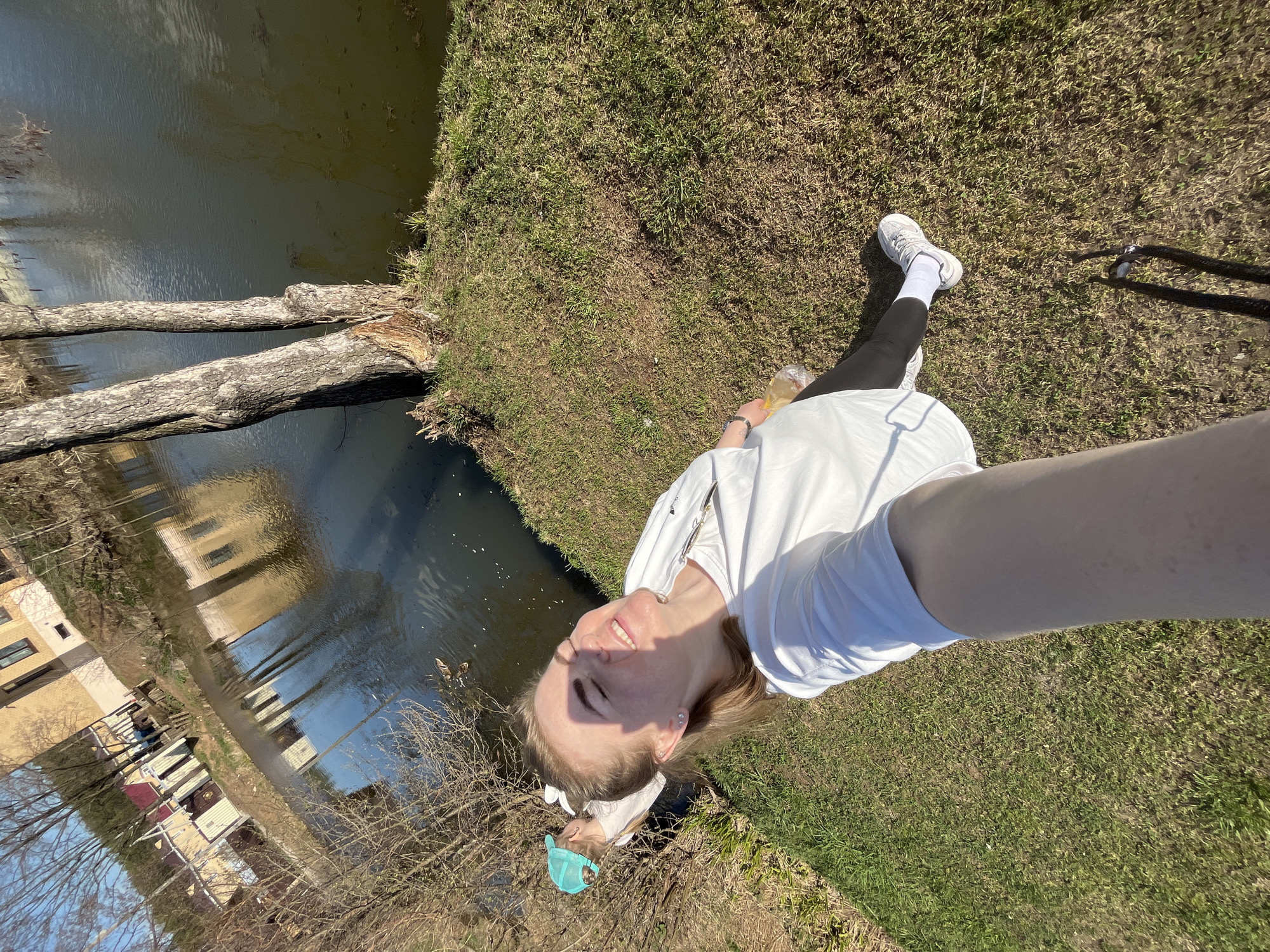 A fair-skinned person is taking a selfie by a body of water. The person is wearing a white t-shirt, black leggings, and white sneakers with white socks. The person is holding a cell phone. There is grass and fallen tree branches beside the body of water. A building is visible in the water’s reflection. A discarded blue face mask is visible on the ground. The location is Chaikovsky, Russia.