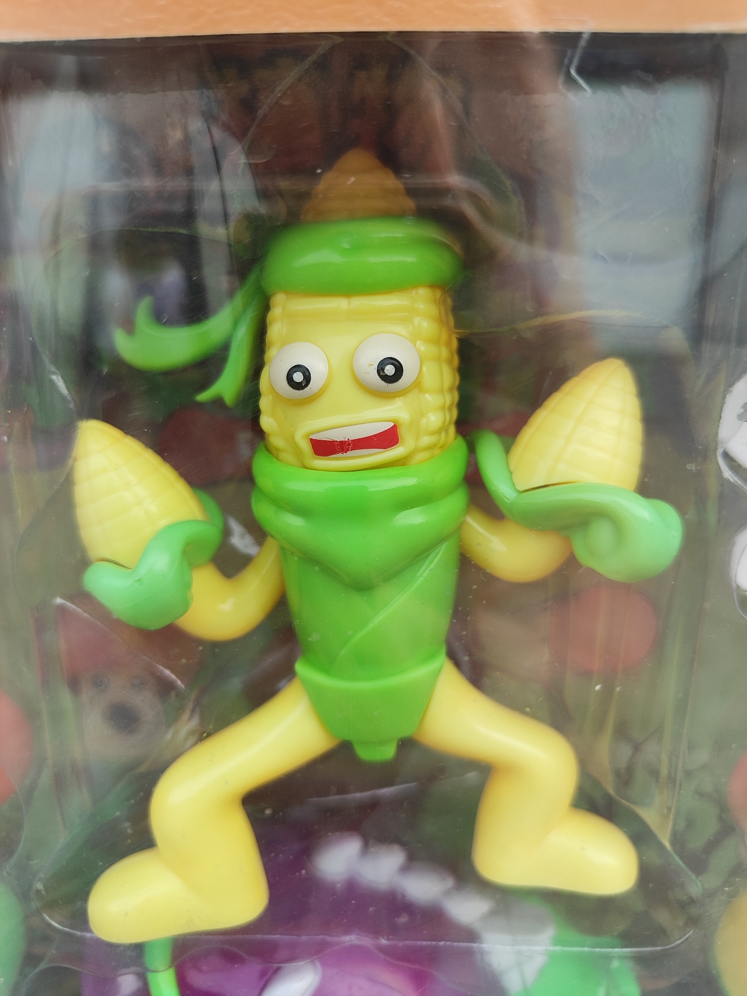 The image shows a close-up of a toy figure of a corn character inside plastic packaging. The toy is yellow and green, with large, cartoonish eyes and a wide mouth. It is holding two mini corn cobs in its hands and has a green scarf-like accessory around its neck and a green bandana on its head. The background is blurred, but appears to be a retail setting. There are no visible text elements in the image. The lighting suggests it is daytime, and there are no indications of weather or specific location cues beyond it being a toy in packaging. The toy has a somewhat surprised or excited expression.