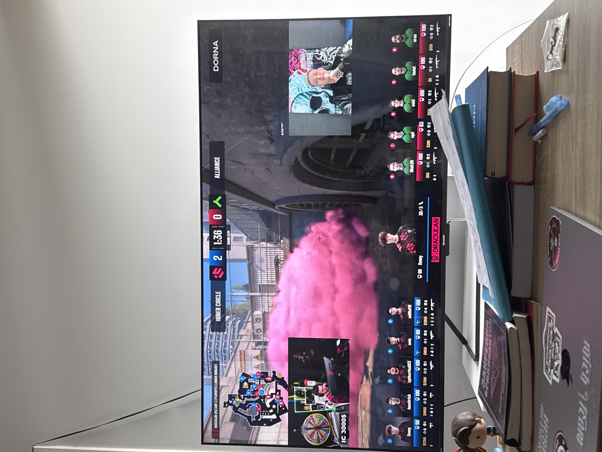 A television screen displays a live gaming broadcast featuring the game "Call of Duty: Modern Warfare II". The broadcast shows a match between "Alliance" and "DORNA" on a map called "Inner Circle". The score is 2-0 in favor of Alliance, with a remaining time of 1:36 in the round. Lower portions of the screen show the maps. Live feeds from two players are in the top corner and bottom center of the screen. Player statistics (names, health, money) for both teams are displayed on either side. The table next to the screen has books, a figure and various smaller items. The model is a Call of Duty eSports match taking place in Warsaw, Poland.