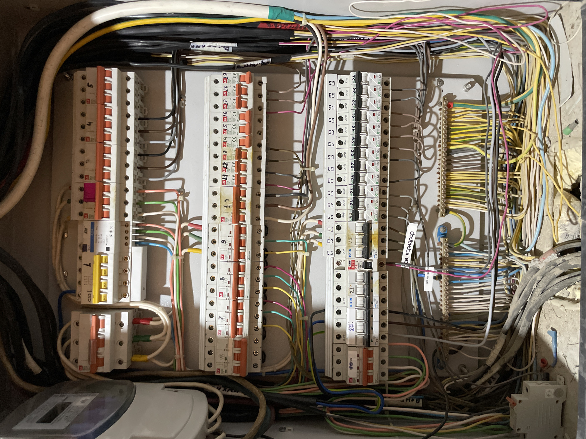 An electrical distribution box is open, revealing a complex arrangement of circuit breakers, wiring, and a meter. The box is mounted on a light gray, textured wall. Several rows of circuit breakers are visible, each with an orange toggle switch. Wires of various colors, including white, yellow, green, red, blue, pink, and gray, are bundled and routed throughout the box. Some wires are connected to the circuit breakers, while others lead to a terminal block at the bottom of the box. A meter, possibly an electricity meter, is attached to the left side of the box, with a gray casing. The environment appears to be an indoor setting, possibly a residential or utility space. The presence of dust and debris suggests the location might be undergoing renovation or is in a less maintained area. This scene is located in Aktau, Kazakhstan.