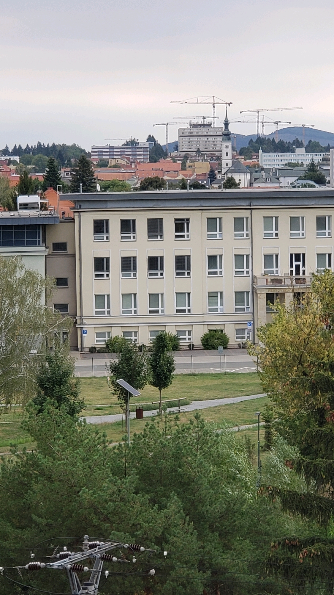 The image shows an aerial view of a city with multiple buildings and construction cranes in the background. The foreground features a light-colored, multi-story building with numerous windows. A green park area with trees and a solar panel is visible in front of this building. In the distance, a skyline of various buildings, including a tall white structure with many windows and a church steeple, is observable. Multiple construction cranes are actively working on different sites within the city. The sky is overcast, suggesting a cloudy day. The scene appears to be a section of Prešov, Slovakia, characterized by its urban landscape and ongoing development.
