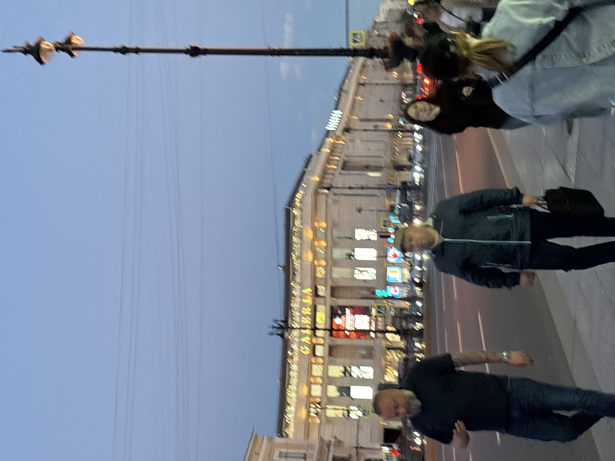 The image depicts a street scene in Saint Petersburg, Russia, at dusk. A large building with illuminated signs, including one reading "GALERIA," is visible in the background, suggesting it is a commercial establishment. Several people are walking on the sidewalk and crossing the street. In the foreground, two men are walking on the sidewalk. The man on the left is wearing a black t-shirt and jeans, and the man on the right is wearing a dark jacket and a cap. A street lamp with multiple lights stands tall on the right side of the image. Cars are visible on the street, indicating ongoing traffic. The sky is a clear blue-grey, typical of twilight.