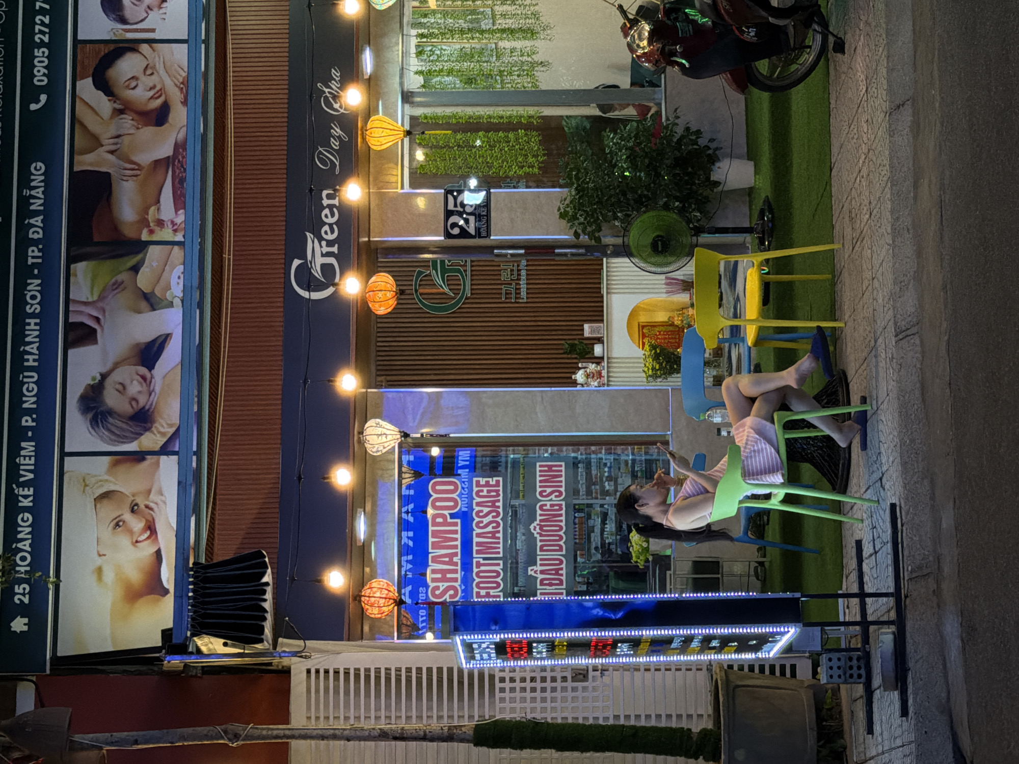 This is a vertical, nighttime, outdoor shot of a street in Đà Nẵng, Vietnam. The scene is dominated by the facade of a business, possibly a spa or salon, with prominent signage. In the foreground, the left side of the image displays a vertical advertisement featuring images of women receiving massages. The text on this advertisement indicates the address "25 HOÀNG KẾ VIÊM - P. NGŨ HÀNH SƠN - TP. ĐÀ NẴNG" and a phone number "0905 272 7-".
To the right of the advertisement, the business's name, "Green Day Cha", is displayed in large, stylized script. Suspended from the ceiling are decorative lanterns, one orange and one white, casting a warm glow. Further into the establishment, a section of the facade displays vertical signage with text including "SHAMPOO", "FOOT MASSAGE", and "ĐẦU DƯỠNG SINH" in Vietnamese.
In the mid-ground, a woman is seated on a green chair, seemingly engrossed in her phone and eating something. She is wearing a pink and white striped dress. Blue and yellow chairs are also visible, suggesting an outdoor seating area. A green oscillating fan stands nearby. Behind the business, a glimpse of the interior shows shelving stocked with various products.
A red motorcycle is parked to the right of the entrance, and the pavement is made of cobblestone. The overall impression is a well-lit commercial street at night, with a focus on services related to relaxation and beauty. The lighting suggests it is evening or night.