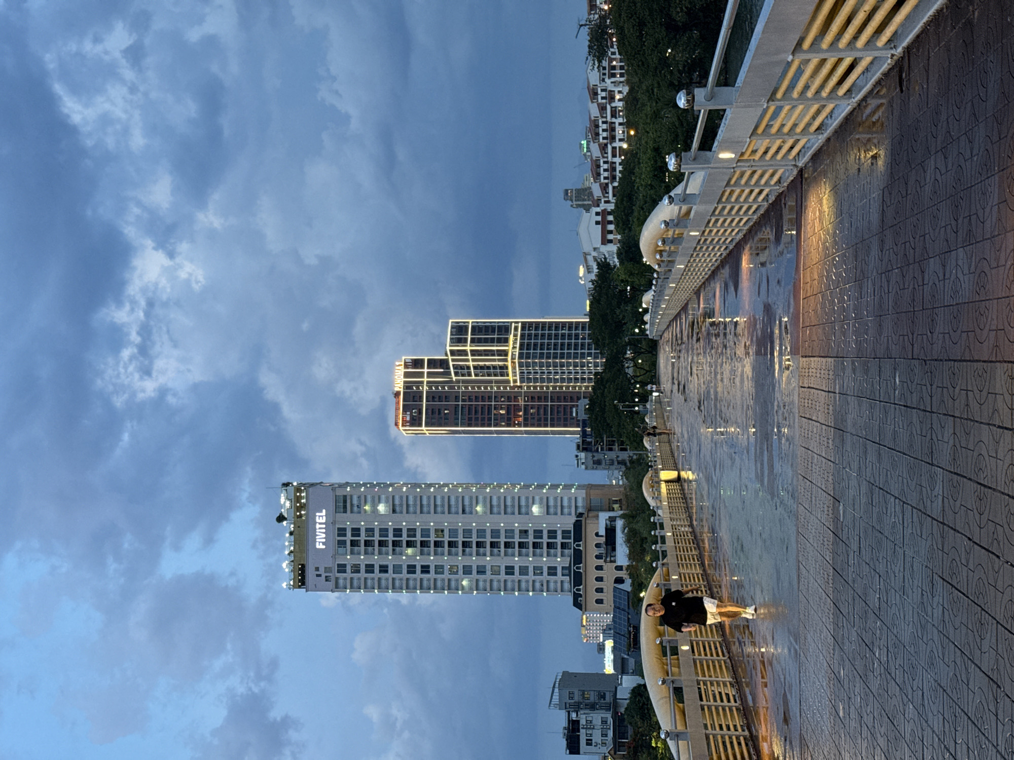 The image depicts a nighttime scene along a waterfront promenade in Đà Nẵng, Vietnam. The perspective is from a raised walkway with a patterned tiled surface that is wet, suggesting recent rain. A metal railing with curved accents and vertical supports lines the edge of the promenade, illuminated by warm-toned lights. Beyond the railing, a body of water reflects the lights from the surrounding cityscape.
In the background, several modern high-rise buildings are visible, some adorned with illuminated windows and decorative lighting. One prominent building prominently displays the text "FIVITEL" in large, bright letters. Another building shows architectural lighting that outlines its structure. Trees and other foliage are interspersed between the buildings, adding a natural element to the urban landscape.
A person, dressed in a black t-shirt and white shorts, is walking along the promenade, their figure silhouetted against the illuminated walkway. The overall mood of the image is calm and serene, with the wet surfaces and evening lights creating a reflective and slightly moody atmosphere. The sky is a deep blue with scattered clouds, indicative of twilight or nighttime.