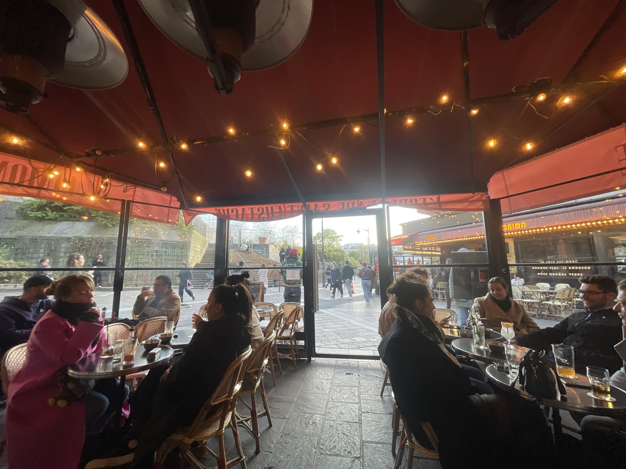 The image depicts an interior view of a covered outdoor cafe terrace located at Place du Tertre in Montmartre, Paris, France. The ceiling is a red fabric awning, from which approximately 15-20 warm-toned string lights are strung, along with two large, silver-colored overhead patio heaters. The floor is composed of dark gray, irregularly shaped paving stones.
Inside the terrace, multiple patrons are seated at round, dark-colored tables with metallic bases, occupying woven rattan chairs with light seat cushions. On the left, a person in a bright pink coat and dark scarf is engaged in conversation, facing to the right. A person in a dark blue top and black cap is partially visible to their left. To the right, a person with dark hair, wearing a dark coat and patterned scarf, is seated with their back mostly to the camera, interacting with a group that includes a person in a light brown coat and a smiling man with glasses, who has a drink in hand. Tables display various beverages in glasses, some appearing to be beer or cocktails, alongside small dishes and a black handbag.
Through the large glass panes and an open central doorway, the exterior environment is visible. On the left, a stone wall with a grid pattern and ascending steps is seen, populated by pedestrians. Through the open doorway, a delivery person wearing a green-backed uniform walks away from the cafe. To the right, a bustling street scene features another cafe or restaurant with a red awning and string lights, bearing prominent signage that includes "A LA MERE CATHERINE." The street is paved, and the scene is illuminated by soft, late afternoon or early evening light, with numerous pedestrians present in the background.