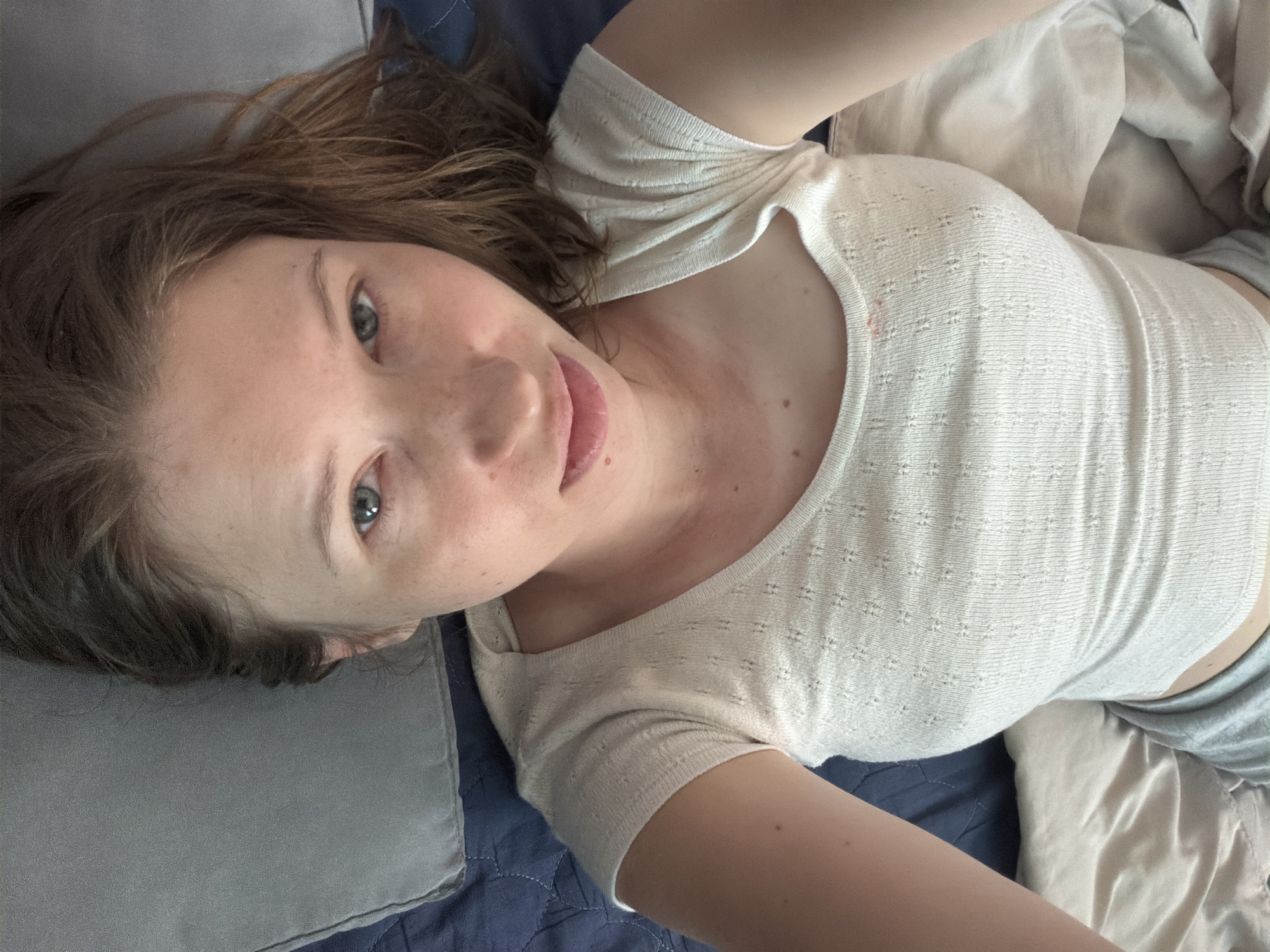 A young Caucasian woman with light brown hair and freckles lies on a bed, looking directly at the camera. She is wearing a light-colored, short-sleeved top. The setting appears to be a bedroom. The image does not contain any information that would allow for a specific location within Dongducheon-si, South Korea.