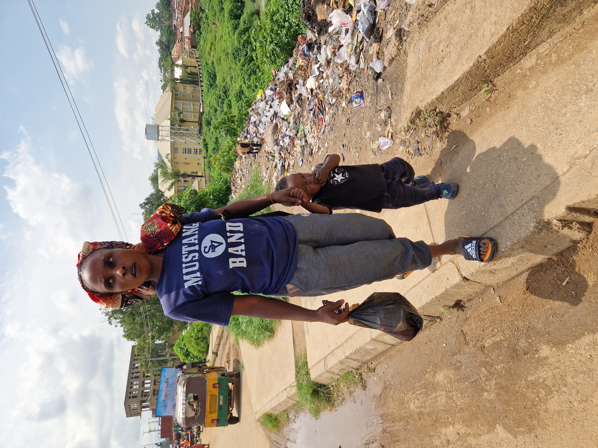An outdoor, eye-level, full shot captures a woman and a young child walking along a dirt and concrete path in Jos, Nigeria. The woman, facing forward and slightly left, wears a blue t-shirt with "MUSTANG BAND" printed on it, gray pants, and a colorful headscarf. She holds a black plastic bag in her right hand. Beside her, the child, also facing forward, wears a black t-shirt with a white star and dark pants, and walks with their left hand held by the woman. To the right of the path, a large pile of garbage covers the ground, extending up a green, vegetated embankment. A brown goat stands amidst the trash. In the background, buildings and trees line the horizon under a partly cloudy sky. A yellow and green auto-rickshaw is visible on the path to the left, along with other distant figures and structures. The scene depicts a daylit environment with a focus on the foreground subjects against a backdrop of urban decay and natural elements.