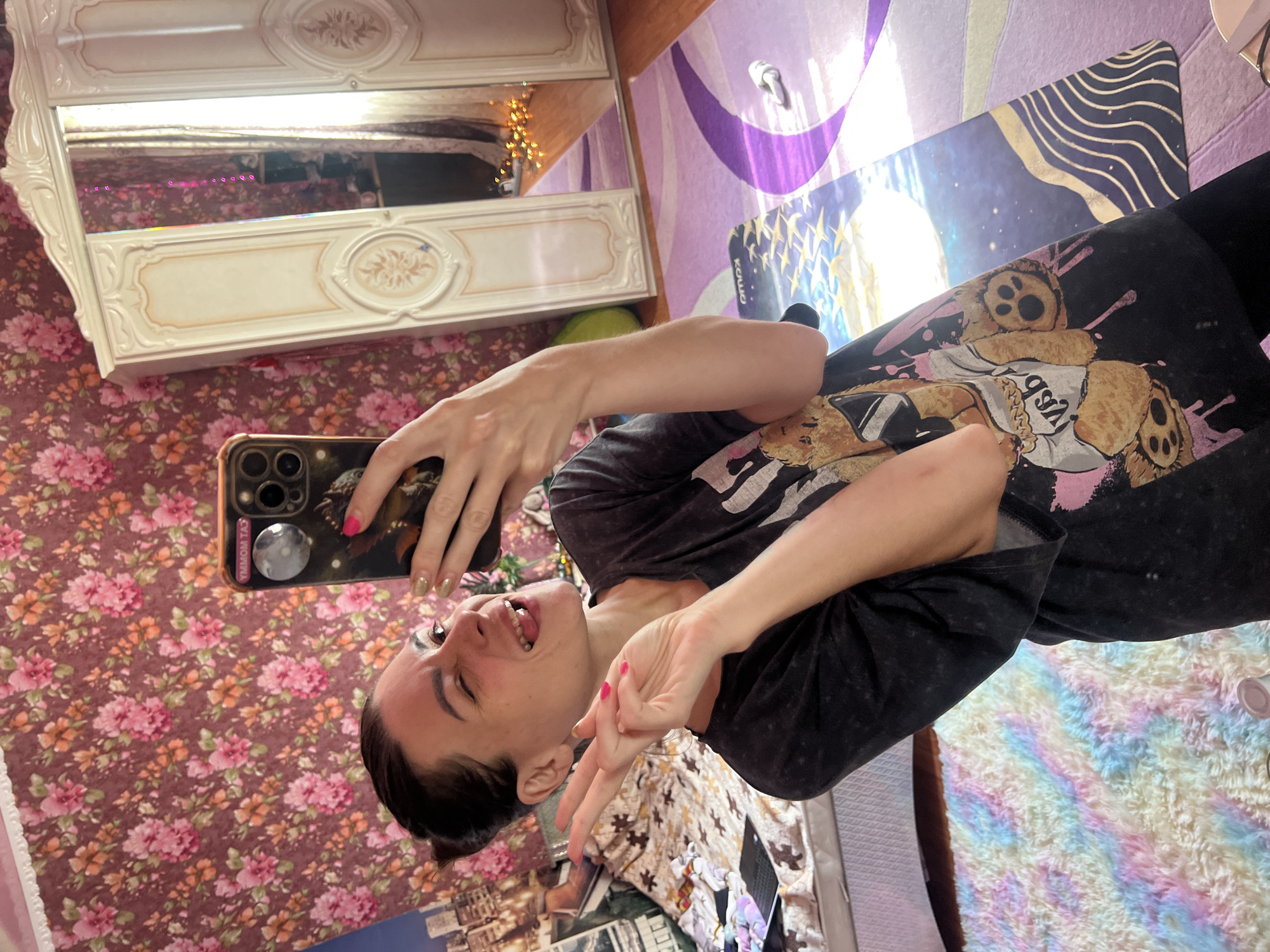 A young woman with dark hair pulled back in a bun is taking a selfie in a room. She is wearing a black t-shirt with a graphic of teddy bears. Her left hand is holding a smartphone, and her right hand is making a peace sign. She has bright pink nail polish on her fingernails and a goldish-green on her thumbs. Her expression is playful, with her tongue sticking out slightly and her eyes closed.
The background features a floral wallpaper with pink and orange flowers. To the left, a white vanity with ornate detailing and a mirror is visible. The mirror reflects the room, showing string lights and possibly a bed. A purple and blue patterned rug is on the floor to the right, along with a black yoga mat with a wavy gold design. The overall impression is that of a brightly decorated, personal space, likely a bedroom. The lighting suggests it could be daytime, but no specific time is indicated. The emotional tone is energetic and fun, captured in the woman's pose and expression. No visible text is discernable on the clothing or other objects, except for text on the phone case which reads "CAT MOM". The location context provided is Krasnoe-na-Volge, Russia.