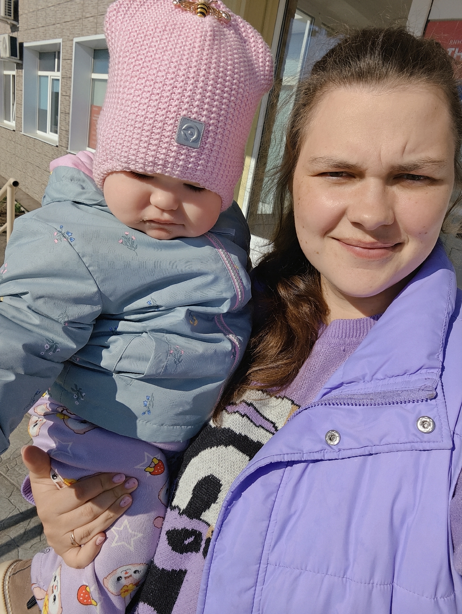 A woman holds a baby in her arms, likely outdoors. The baby wears a pink knit hat with a bee embellishment, a blue-grey jacket patterned with small floral designs, and patterned light purple pants with images of strawberries, stars and a cartoon animal. The woman wears a purple sweater and a light purple puffer vest. The location shows a building of light stone and windows to the left, and possibly an entranceway to the right. The scene is set in Chaikovsky, Russia.