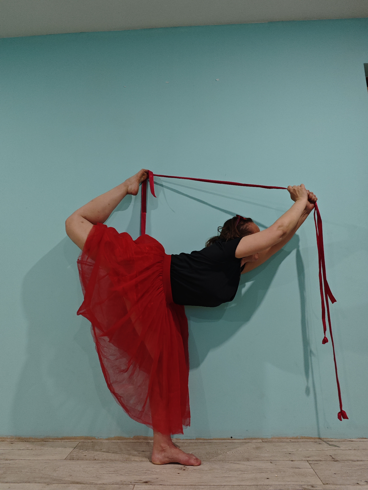 A woman in a black top and red tulle skirt is captured mid-yoga pose against a light blue wall. She is holding a red ribbon that loops around her right foot. The setting appears to be an indoor space with a light-colored wooden floor. The location, Markovo, Russia, cannot be verified from the visual data.