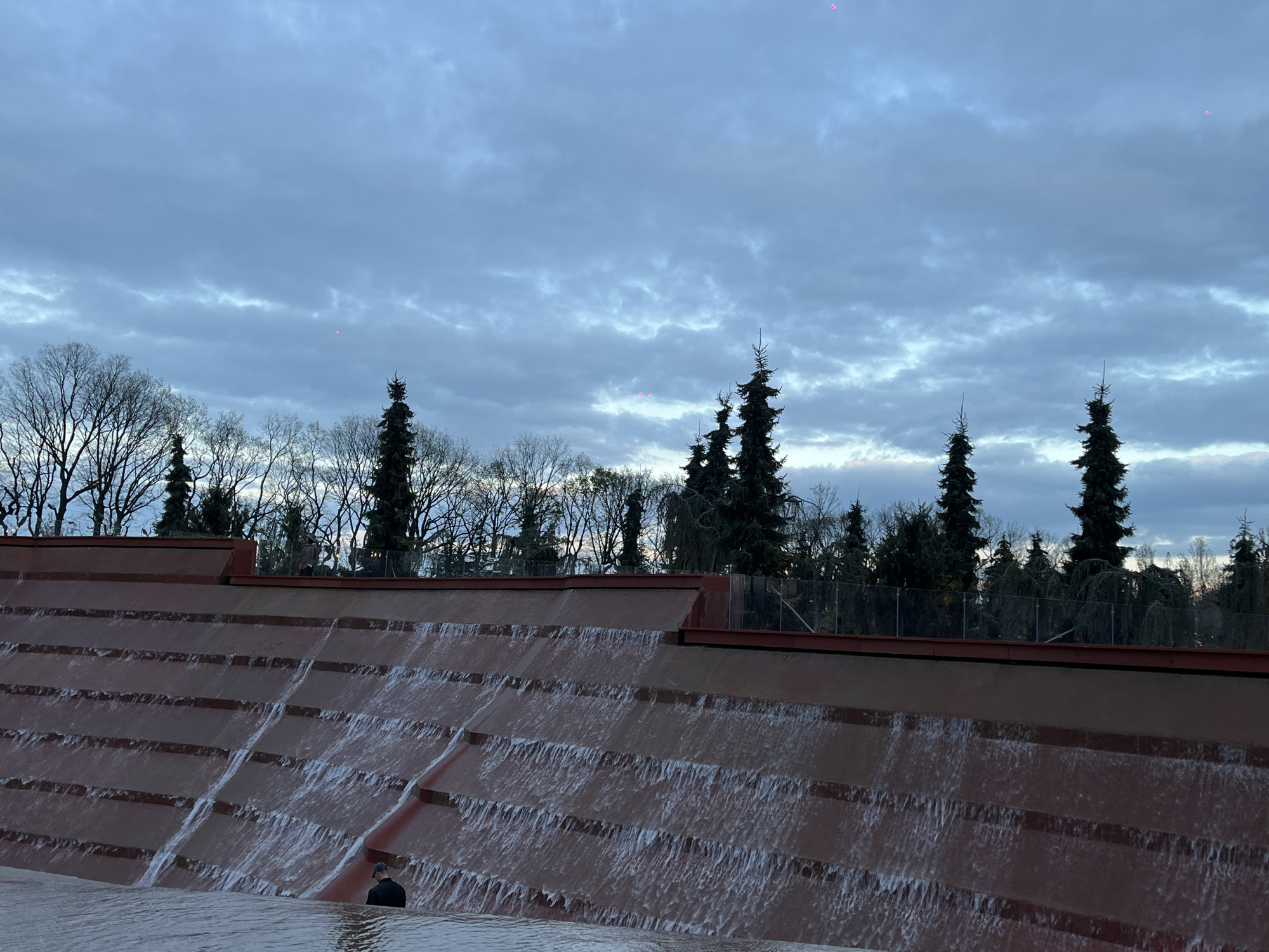 This image depicts a large, terraced artificial waterfall, characteristic of a modern park or public landscape in Krasnodar, Russia. The waterfall structure is constructed from reddish-brown material, with water cascading down multiple broad steps.
In the lower right foreground, a person wearing a dark jacket and cap is partially submerged in the pool at the base of the waterfall, suggesting they are wading or performing maintenance. Above the waterfall, behind a low glass barrier, another person is faintly visible, standing on what appears to be a walkway or viewing platform.
The background features a dense line of trees, comprising both bare deciduous branches and dark evergreen conifers. Above the tree line, the sky is overcast with thick, grey-blue clouds, indicating a cloudy day or approaching twilight. Several small, bright pink dots, likely lens flares or sensor artifacts, are noticeable against the cloudy sky. The scene conveys a calm, somewhat muted atmosphere due to the soft lighting and cloudy conditions. No discernible text is visible in the image.
