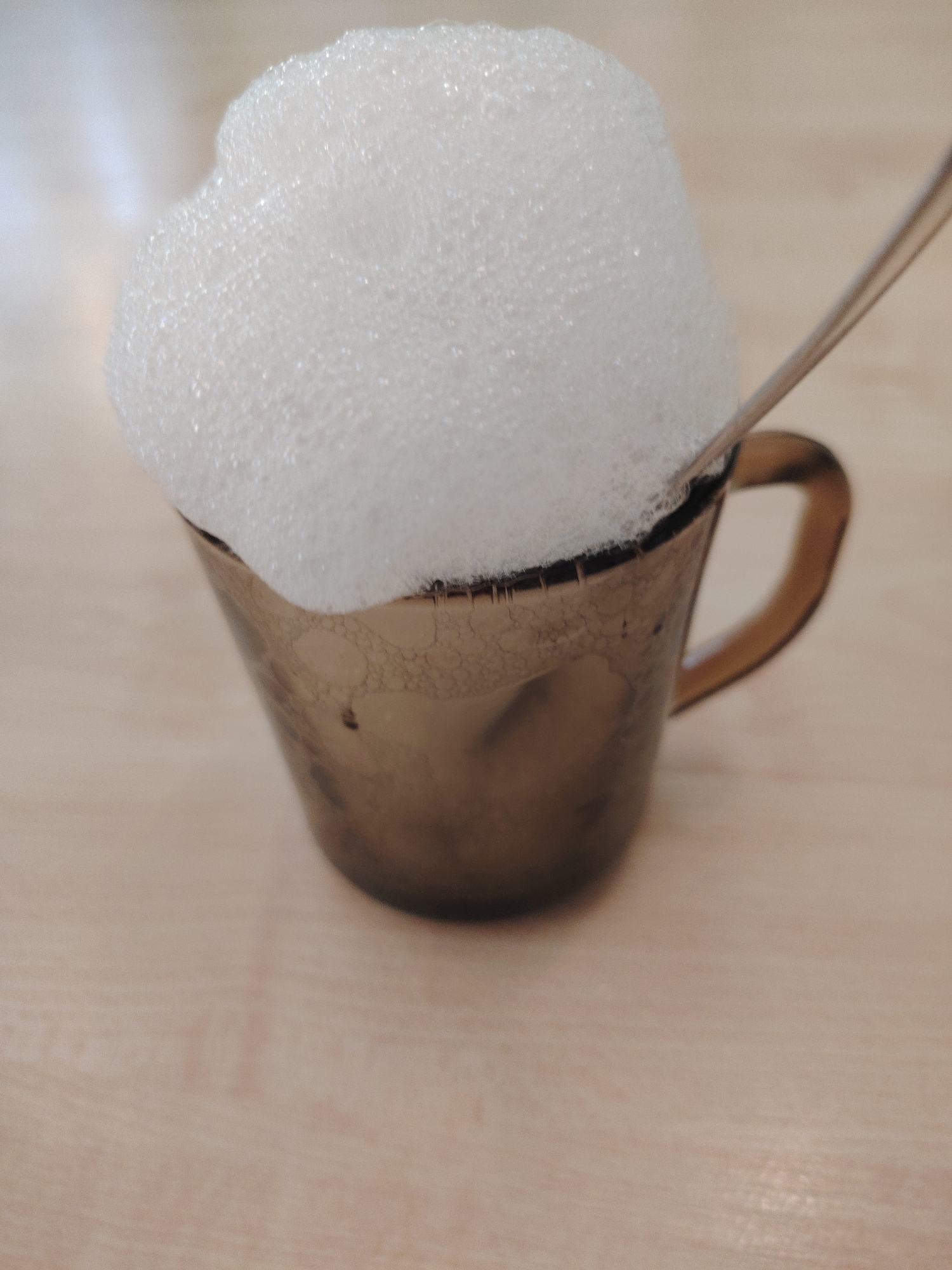 A close-up shot depicts a beige-colored mug filled to overflowing with a large mound of white foam. A metallic spoon is partially visible, inserted into the foam. The mug rests on a light-colored, possibly wooden, surface. No people are visible. The location of the image is not discernable.