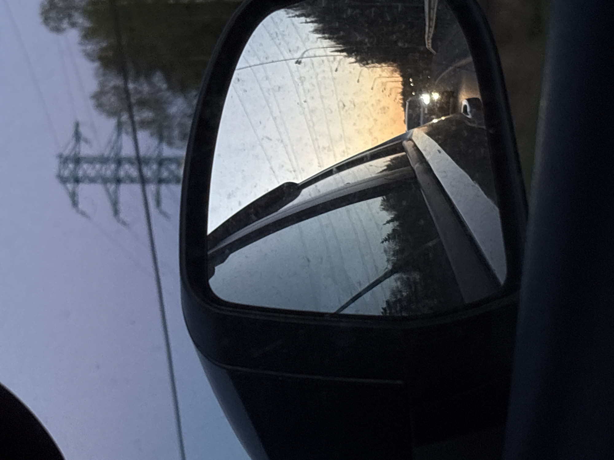 The image depicts a car's side mirror reflecting a road and surrounding environment. The mirror shows a roadway with at least one other vehicle with headlights visible in the distance. Trees are visible along the side of the road in the mirror. A utility pole is visible on the left side of the image, out of the mirror's reflection. The image appears to have been taken at dusk or dawn given the lighting. The location is inferred from the prompt as Myaglovo, Russia. There are no people visible in the image.