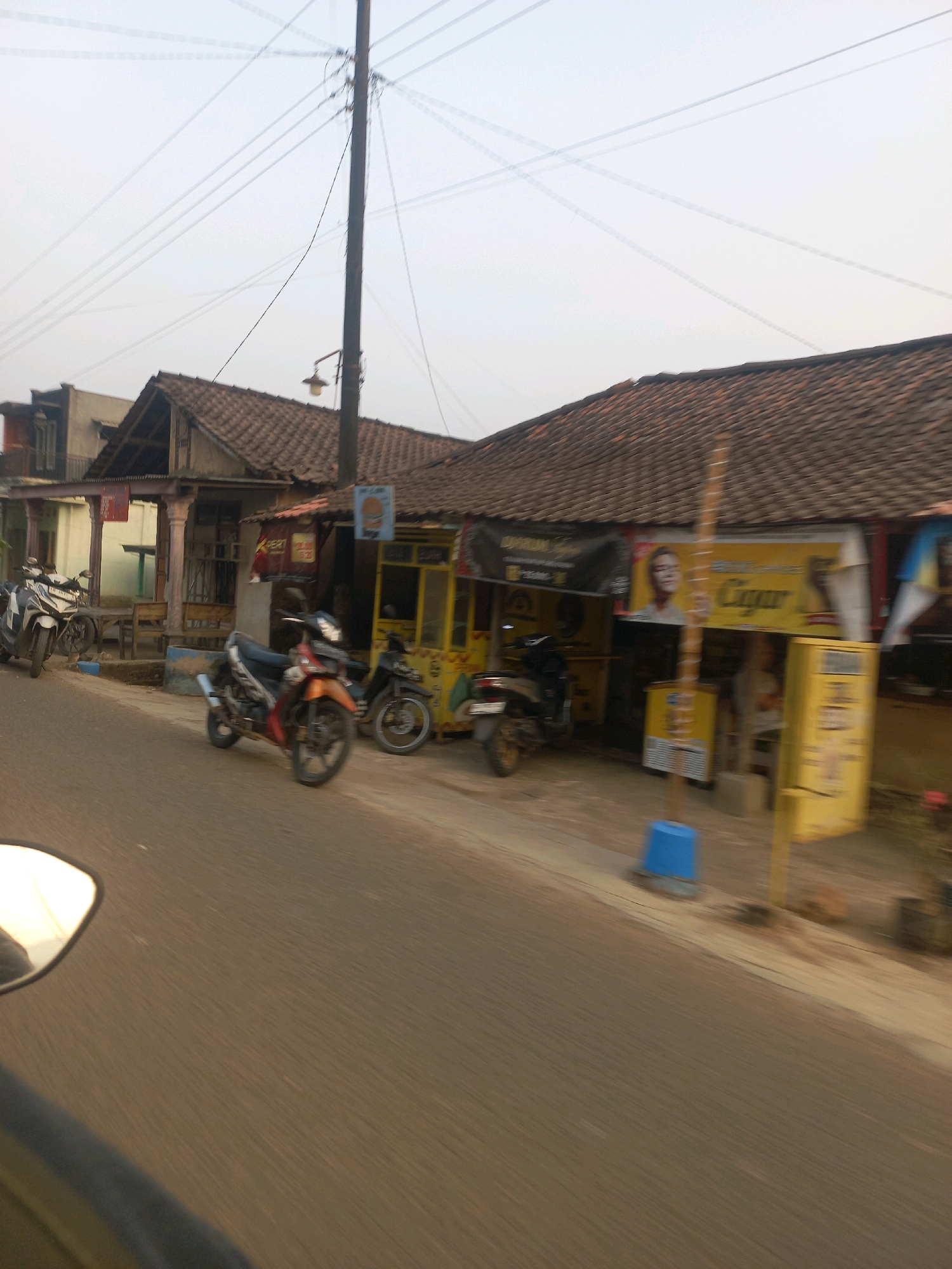 The image depicts a street scene in Sukoharjo, Indonesia. A road runs horizontally across the foreground, with the edge of a vehicle's side mirror visible in the lower left corner, reflecting a bright sky. On the right side of the road, a row of buildings with tiled roofs are visible. These buildings appear to be commercial establishments. One building has a yellow facade with signage that includes the word "Cigar" and a portrait of a person. Another building has a dark awning with text. Several motorcycles are parked along the road and in front of the buildings. One motorcycle, orange and black, is prominent in the center, and another white and black scooter is parked further to the left. A utility pole with electrical wires stretches vertically in the background. The overall atmosphere suggests a typical daytime street view in a developed area of Sukoharjo.