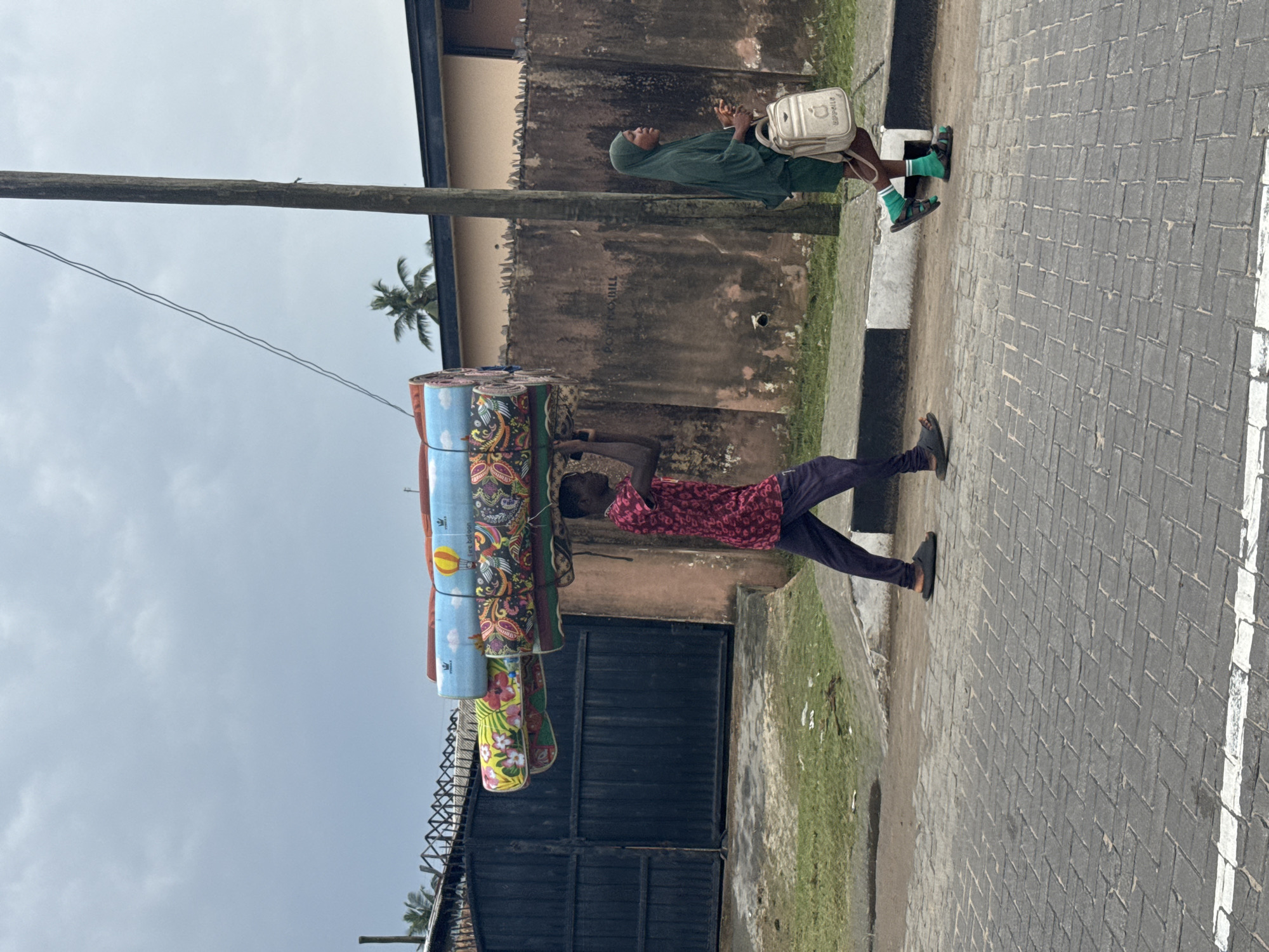 A man carries a stack of rolled mats suspended from an overhead pole in Badagry, Nigeria. He wears a red patterned shirt and dark pants. A woman wearing a green hijab sits on the curb nearby with a white bag. The scene is set on a paved street with a white line marking, adjacent to a building with a gated entrance and a weathered wall. An electrical pole is visible overhead. The sky is overcast.