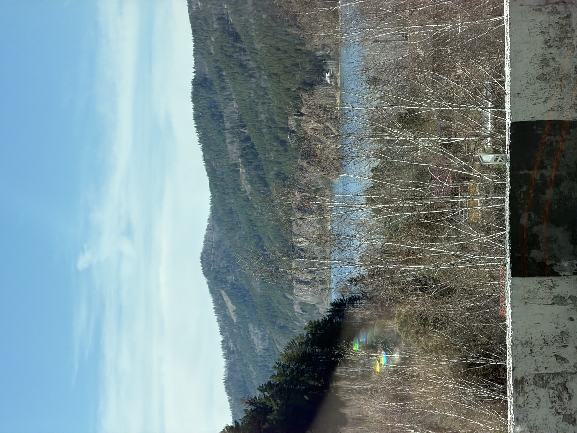 The image shows a landscape with a body of water, a tree-covered mountain, and a partially visible white wall or structure in the foreground. There is a partially cloudy blue sky. The bare branches of trees are interspersed between the wall and the view. Rainbow flares are visible within the frame, likely caused by light refracting through the lens. There are no visible people. The location appears to be rural. The image suggests that the view is being captured from inside a vehicle due to the presence of the wall. The landscape likely corresponds to the city of Слизнево, Russia.
