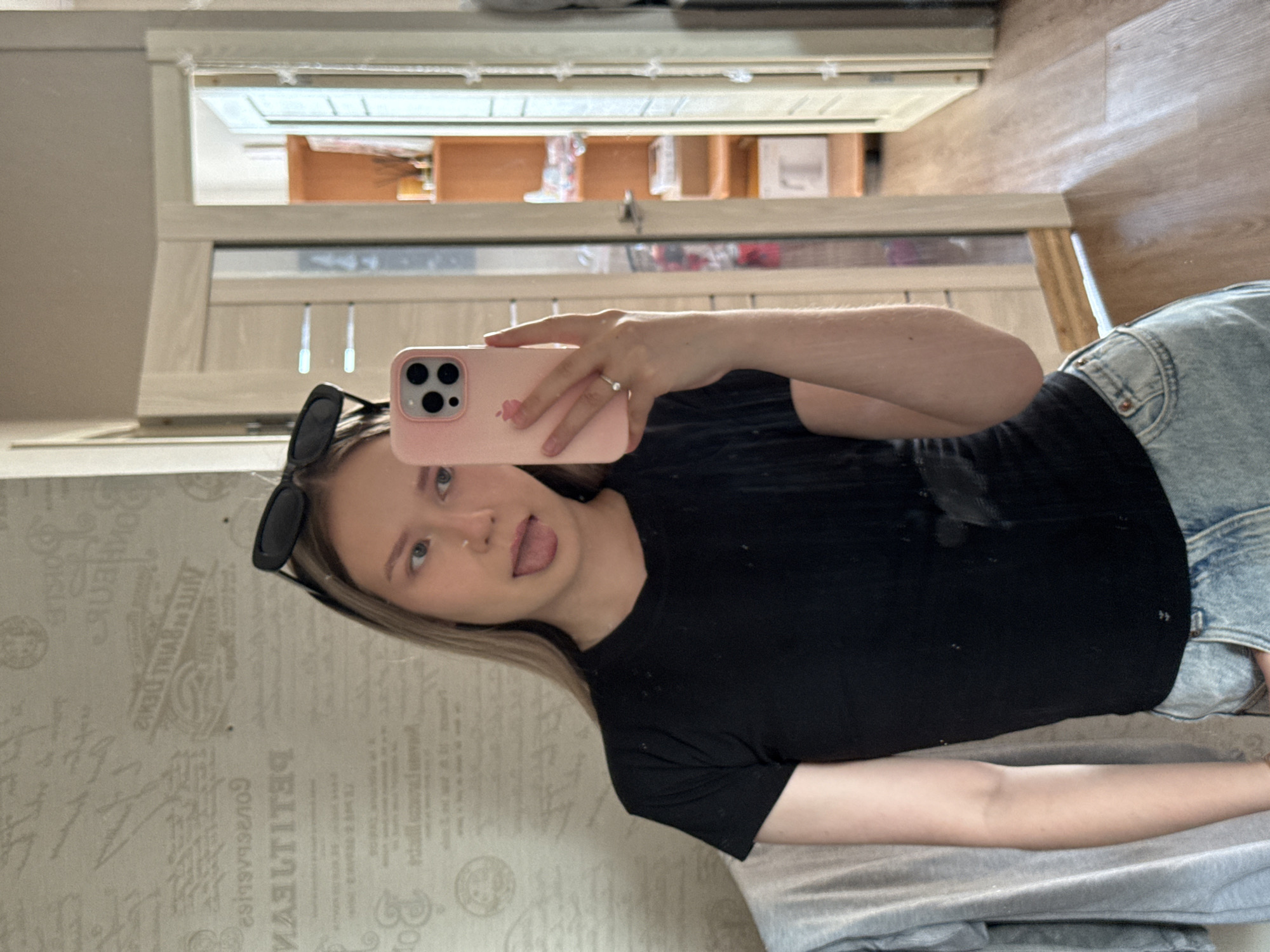 A woman is taking a selfie in a room. She is wearing a black t-shirt and blue jeans and has blonde hair. She is holding a pink smartphone. The background is a room with patterned wallpaper. The location is unknown.