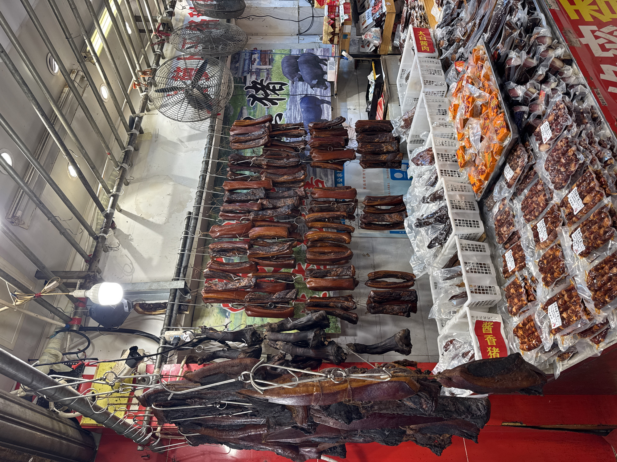 This image depicts a meat market stall in Qingyang District, China, featuring a wide assortment of cured and smoked meats. Suspended from metal racks are numerous strings of various cuts of meat, some appearing dark and aged, while others are lighter brown and possibly smoked or dried. The meats vary in size and shape, including what appear to be hams, sausages, and other processed meat products.
In the foreground, a display table showcases pre-packaged meats in clear plastic wrapping. These packages contain smaller, uniform pieces of cured meat, likely sausages and possibly sliced bacon or jerky. A large metal tray in the center holds pieces of cured meat cut into square or rectangular shapes, also packaged. White plastic baskets are visible behind these packaged goods, some containing more unwrapped cured meat products.
The background reveals a wall with a poster featuring pigs and Chinese characters, suggesting the type of products sold. Two large industrial fans are mounted above the hanging meats, likely for ventilation and drying. Lighting fixtures are visible on the ceiling, illuminating the market stall. The overall environment appears to be an indoor market, with metal shelving, exposed pipes, and a red-colored partition on the left side. No people are actively visible in the frame.
