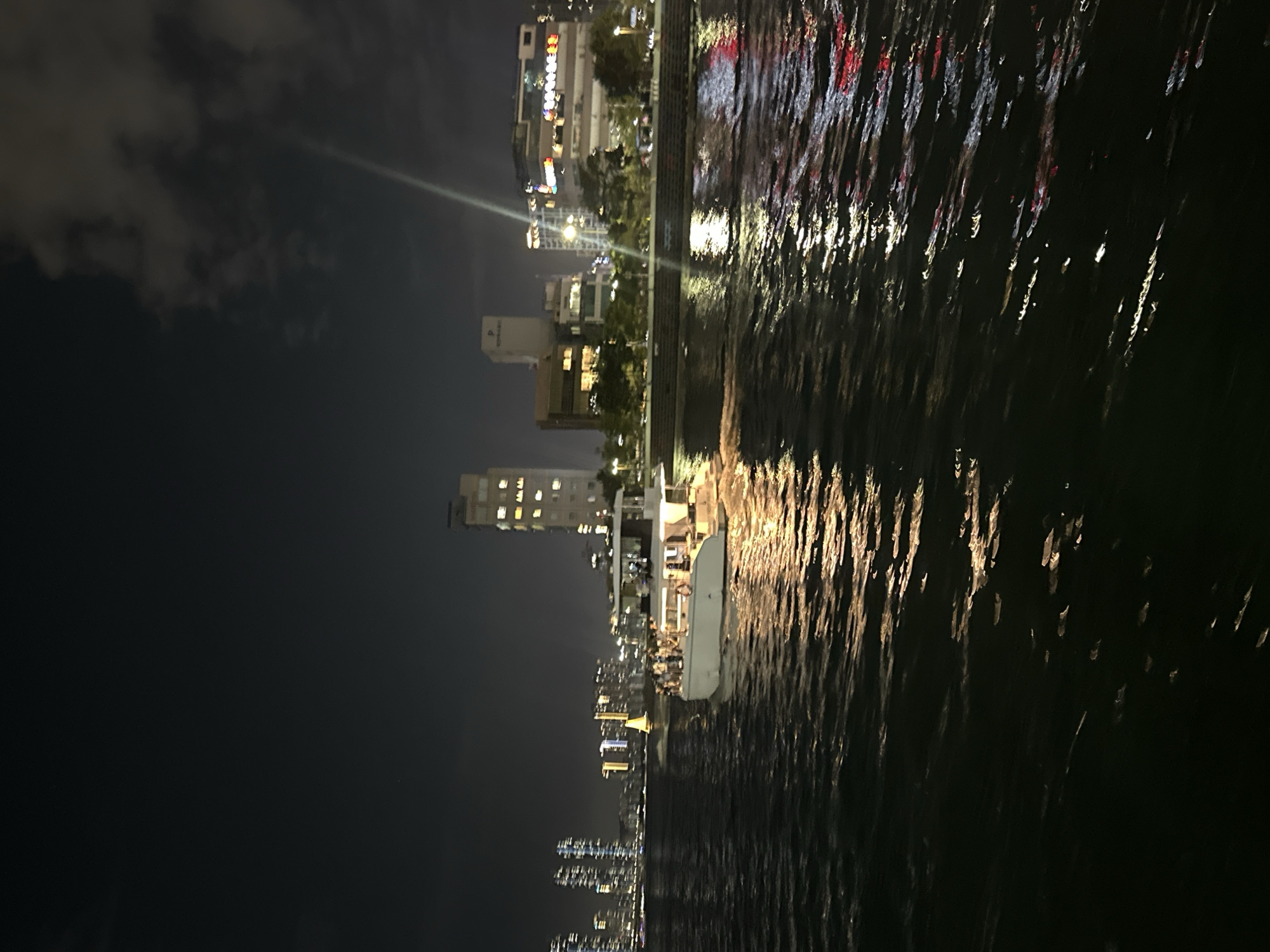 A night-time scene captures a wide body of water, likely a river or bay, with the Busan skyline visible in the background. The water's surface is rippled, reflecting the lights from the city and a brightly lit white boat in the foreground. The boat, which appears to be a passenger vessel, has several people standing on its deck, suggesting a tour or event. In the distance, numerous tall buildings, illuminated with lights, form a dense urban landscape. A yellow sail boat is also visible further out on the water. The sky is dark, with clouds and a faint light beam extending from the upper right. The scene conveys a sense of urban nightlife and recreation on the water.