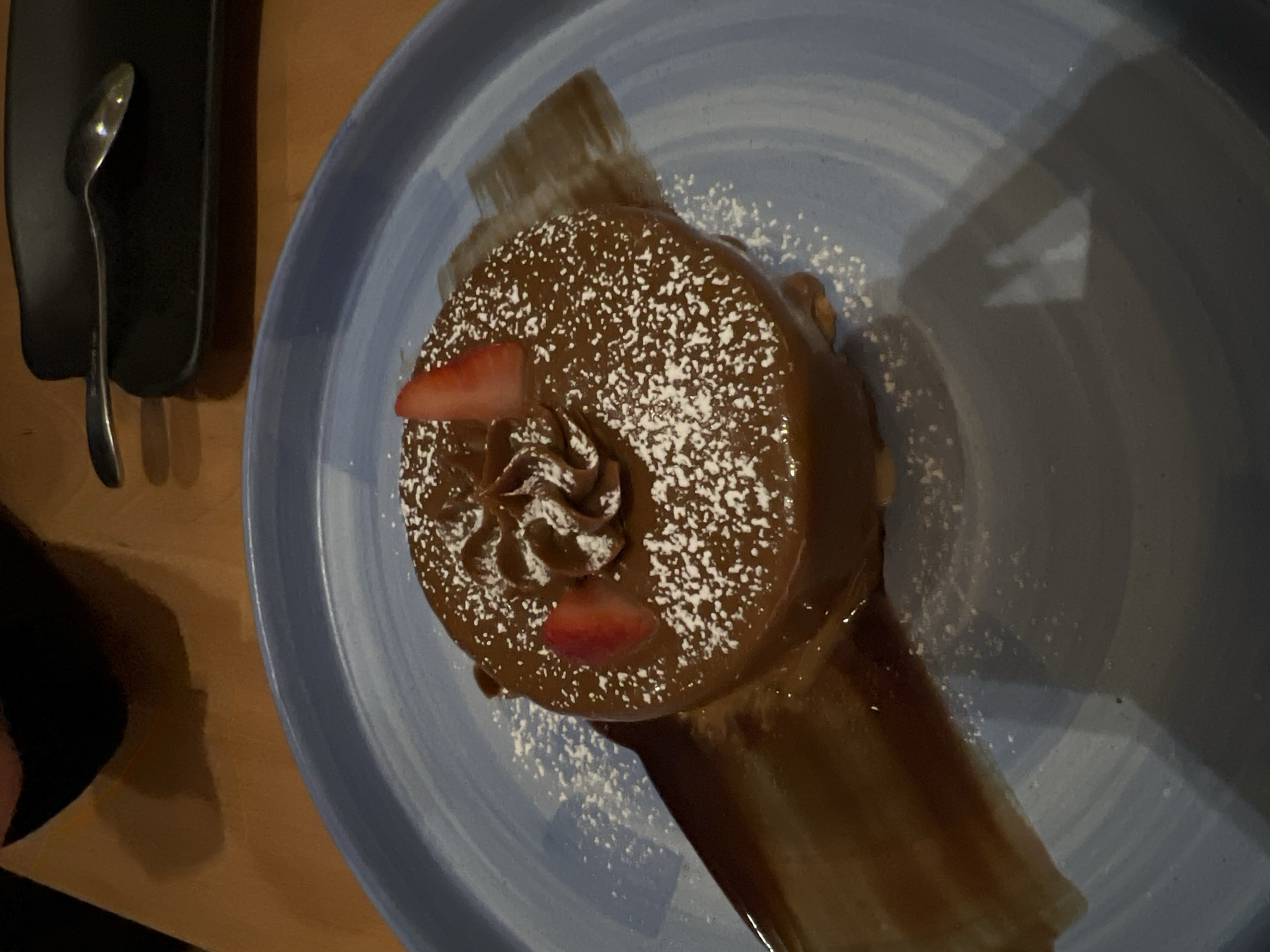 The image displays a close-up, top-down view of a chocolate dessert served on a light blue ceramic plate, likely in an indoor setting such as a restaurant or cafe in Mexico City, Mexico.
The main subject is a round chocolate dessert, possibly a cake or a rich mousse, generously coated in a smooth, glossy chocolate ganache or frosting. Its surface is dusted with white powdered sugar, creating a delicate speckled pattern. Two bright red strawberry slices, acting as a garnish, are placed on top of the dessert: one near the upper edge and another towards the lower right. A swirl of what appears to be chocolate cream or frosting is piped in the center of the dessert. Beneath the dessert, a rich chocolate sauce is artfully spread across the plate.
To the left of the plate, a metallic spoon rests on a dark, rectangular surface, suggesting it is prepared for the dessert's consumption. The background features a warm, wooden table surface, with shadows indicating a dimly lit environment, possibly an evening setting. No people are visible, and no discernible text is present in the image. The overall scene emphasizes an appealing and carefully presented dessert.