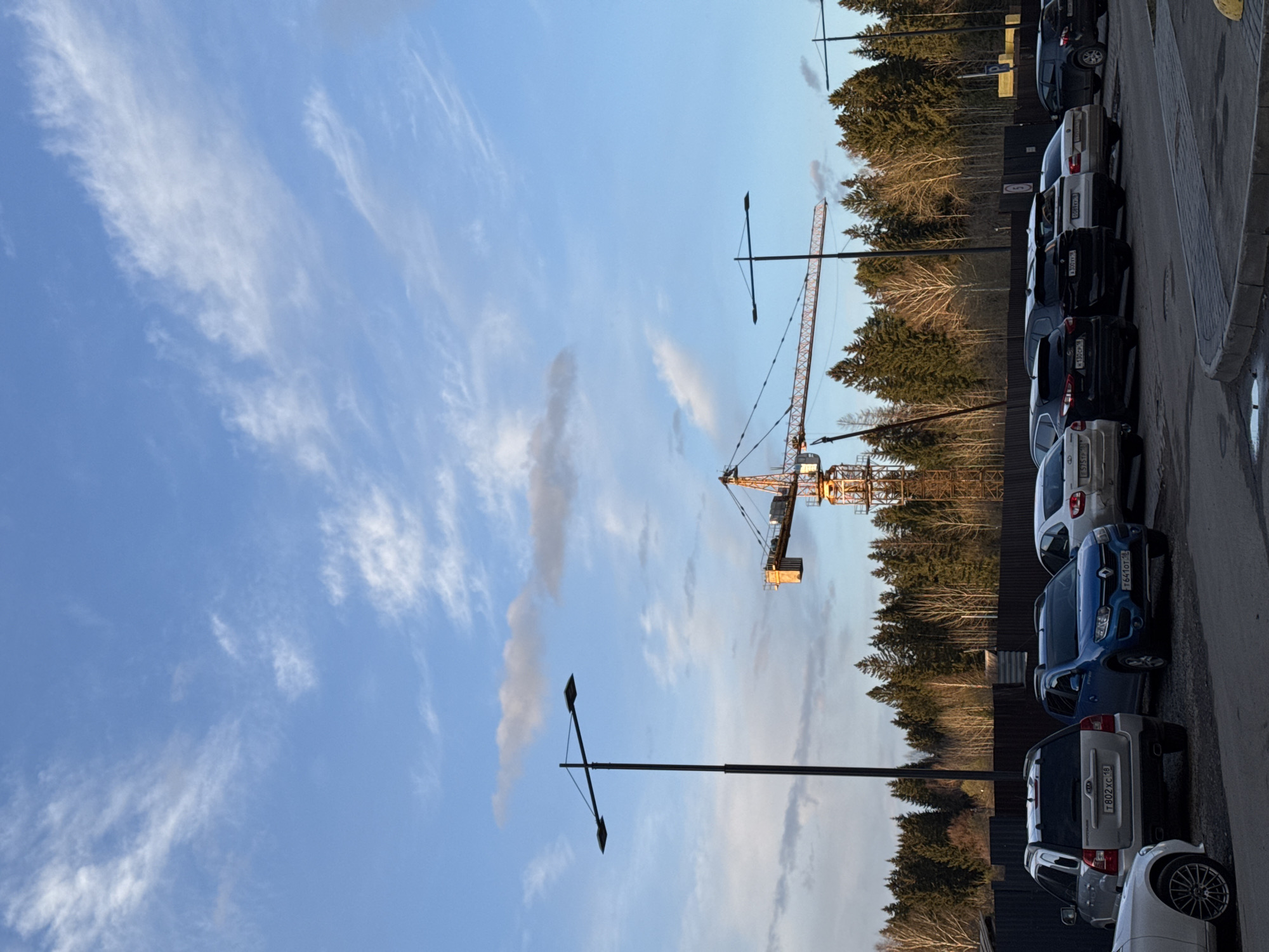 This horizontally oriented image captures a roadside scene in Izhevsk, Russia, featuring a parking area, a forest, and a construction crane under a clear sky.
In the foreground on the right, a line of cars is parked along a paved curb. Visible vehicles include a white car with license plate E575PC 18, a black car with K132CP 18, a blue car with T641OT 18, a silver SUV with T802XC 18, a white car with B307YY 18, and another dark car with A930XH 18. All visible license plates bear the region code "18," corresponding to the Udmurt Republic, where Izhevsk is located.
Parallel to the parking area, a dark wooden fence separates the road from a dense forest of evergreen trees. Some deciduous trees with bare branches are also visible within the forest. In the mid-ground, a prominent yellow and black tower crane stands tall, indicating a construction site. Two black streetlights, featuring horizontal crossbars with square light fixtures, are positioned at different distances in the scene. A circular speed limit sign, displaying the number '5', is partially visible on the wooden fence.
The sky is a bright blue, adorned with scattered, wispy white clouds. The lighting suggests it is either late afternoon or early evening, with a soft, warm glow on the landscape. No people are visible, and no specific activity is taking place, presenting a static blend of urban infrastructure, natural forest, and industrial development.