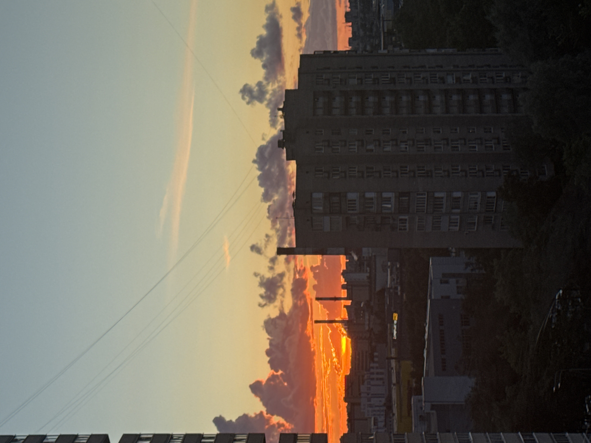 An outdoor, high-angle, wide shot captures a city skyline at sunset. The sky transitions from a light blue at the top to a vibrant orange and yellow near the horizon, where a large, fiery sun is setting behind industrial buildings and apartment complexes. Clouds, illuminated by the setting sun, are scattered across the sky, with some appearing dark and ominous. Several tall, thin industrial smokestacks are visible in the midground, emitting wisps of smoke. In the foreground, a large, multi-story apartment building dominates the right side of the frame, its facade featuring numerous windows and balconies. Another, similar building is partially visible on the far left. Trees are visible in the lower portion of the image, obscuring some of the ground level. Thin, linear structures, possibly wires, are stretched across the sky in a diagonal pattern. The overall impression is of an urban environment at dusk, with the dramatic colors of the sunset contrasting with the more muted tones of the buildings and industrial structures. The scene is devoid of people or any overt activity, presenting a static view of the city.