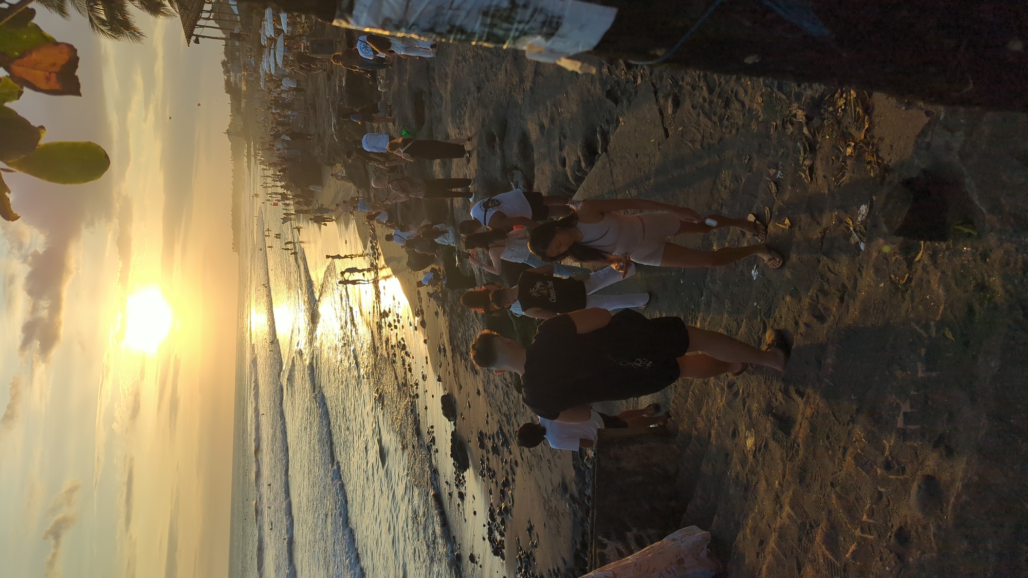 The image depicts a crowded beach scene during sunset in North Kuta, Indonesia. The sun is low on the horizon, casting a warm golden glow across the sky and ocean, with reflections shimmering on the water's surface. Waves gently break on the sandy shore, which is dotted with rocks and debris. A large number of people are present, spread across the beach and along the rocky shoreline.
In the foreground, a man wearing a black t-shirt and shorts stands facing away from the camera, with his back to the ocean. To his left, a young boy in a white t-shirt and shorts is also facing away. To the right of the man in the foreground, a woman in a light-colored sleeveless top and shorts is looking down at her phone. Further into the scene, other individuals and groups are visible, some walking, some standing, and some interacting with each other.
The background shows a long stretch of coastline with more people, buildings, and palm trees. A few umbrellas are set up on the beach, suggesting a commercial area. The overall atmosphere appears to be one of leisure and social gathering, likely a popular spot for sunset viewing. The lighting suggests it is either sunrise or sunset, with the golden hour dominating the scene.