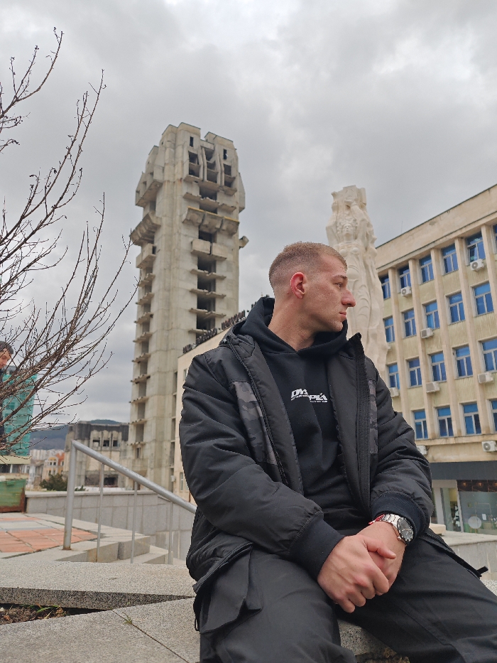 The image displays a young adult male seated on a concrete surface in an urban plaza in Shumen, Bulgaria, under an overcast sky. He is positioned in the foreground, facing right in profile, with his hands clasped. He wears a black padded jacket, a black hooded sweatshirt with white text "DVDRPLA" on the chest, and black cargo pants. A silver-toned wristwatch with a red accent is visible on his left wrist. To the far left, partially obscured by bare tree branches, another individual's head and shoulder are visible.
In the background, a tall, unfinished concrete brutalist-style structure, identifiable as the "House of Culture," dominates the left side. To the right, a large, light-colored abstract monumental sculpture, part of the "Monument to 1300 Years of Bulgaria," is visible behind the main subject. Adjacent to the monument, a multi-story building with a light-colored facade, numerous blue-framed windows, and exterior air conditioning units, consistent with the Shumen Municipality building, is present. The immediate foreground features bare tree branches and a concrete paved area with visible steps and a metal handrail. The sky is a uniform light grey, indicating an overcast condition.