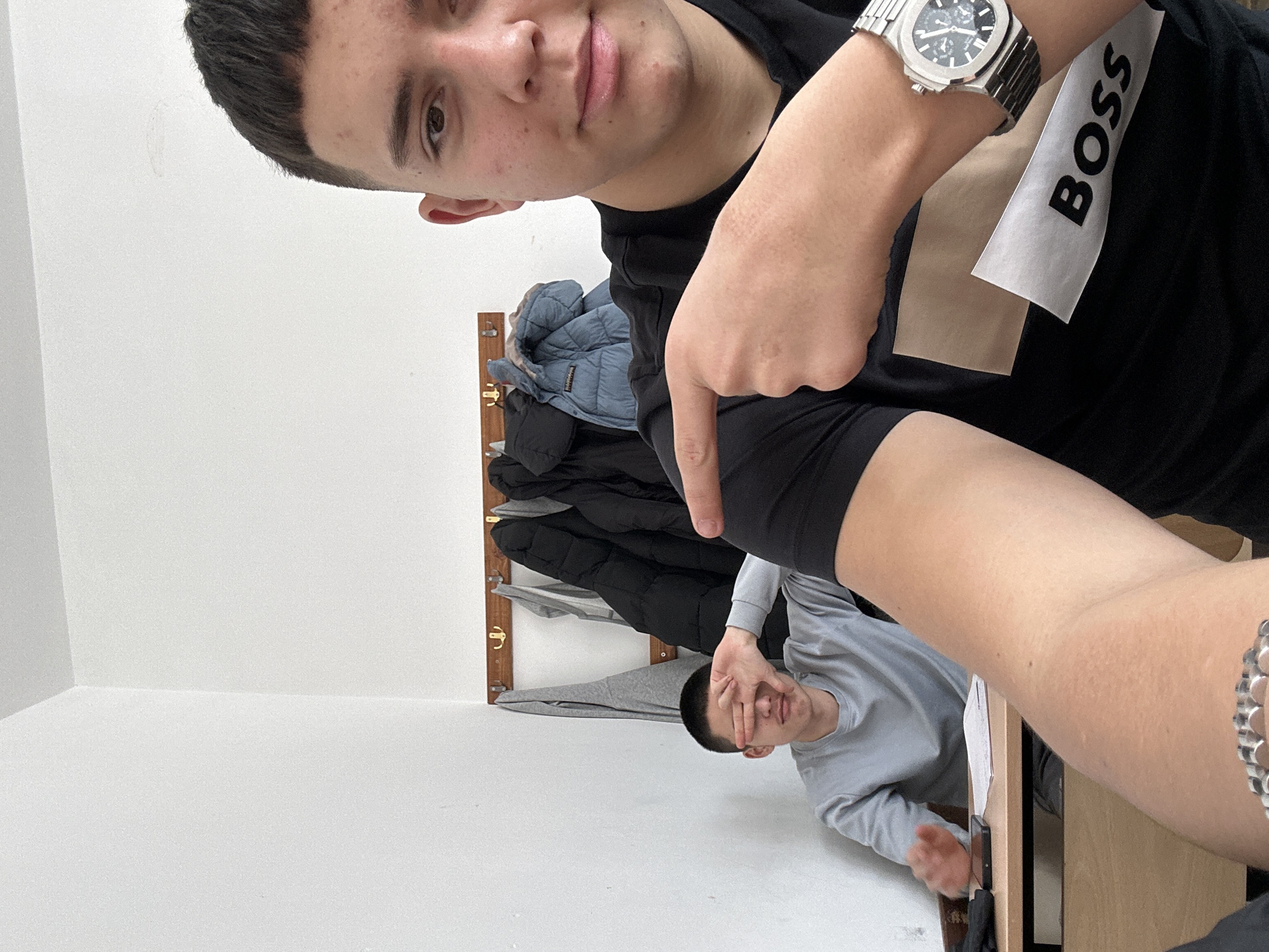 The photograph shows two young men in an indoor setting, likely a small office or classroom.
The first man is in the foreground, partially obscured by the camera angle. He has short dark hair and is wearing a black t-shirt with the word "BOSS" visible on the chest. He is pointing downward with his right hand. A silver wristwatch is visible on his left wrist.
The second man is seated behind a desk, wearing a light grey long-sleeved shirt. He is covering his face with his left hand and appears to be smiling slightly.
In the background, a light-colored wall is visible, along with a coat rack holding several jackets and a grey garment. The overall lighting is bright.
The location is not definitively known to be Zlatograd, Bulgaria from the image itself.