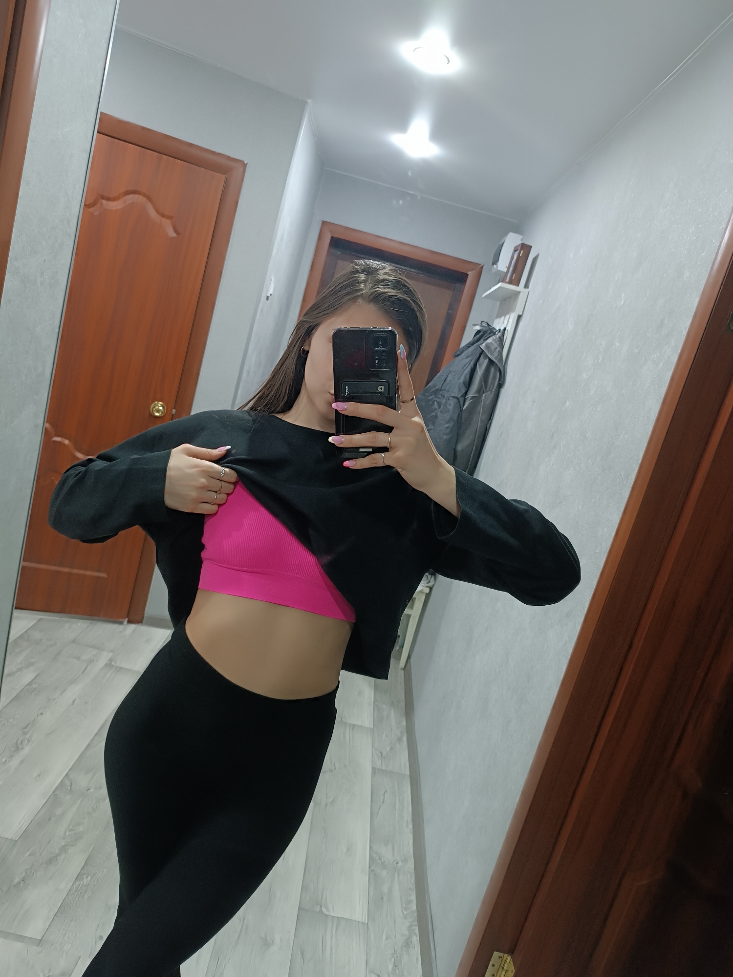 A woman stands in an indoor hallway taking a selfie in a mirror. She is wearing black leggings and a bright pink sports bra under a black long-sleeved shirt, which she is partially lifting. The hallway has two brown doors and light gray walls, with a coat hanging on a rack in the background. There is a ceiling light visible, and the mirror reflects the hallway's layout. The location is Unknown, Unknown.