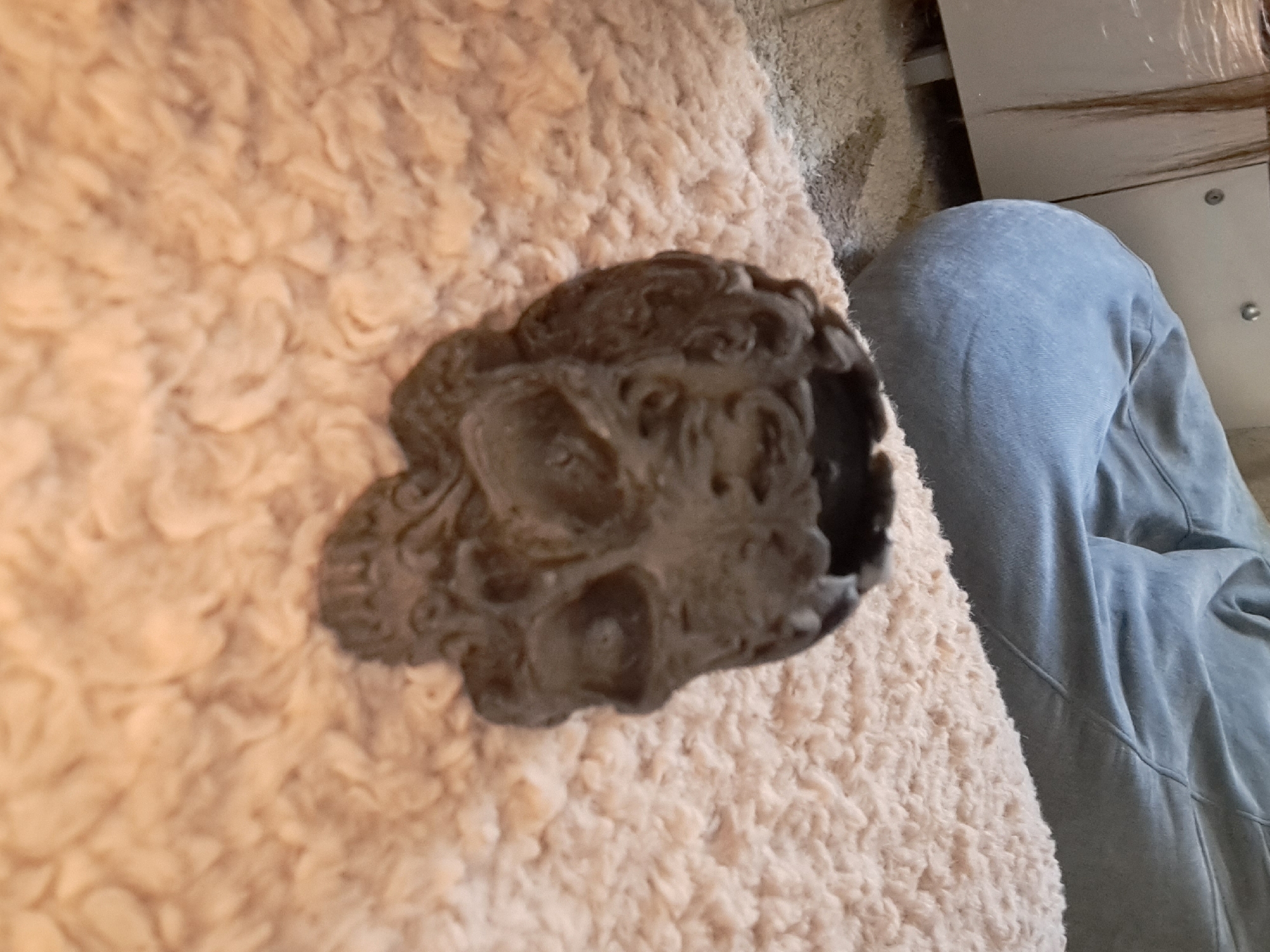 A skull-shaped planter sits on a fuzzy peach-colored rug. Behind the planter, a light blue beanbag chair is partially visible. To the left of the beanbag chair is a white cabinet. The scene appears to be indoors, possibly in a living room or bedroom. The overall lighting is dim. There are no people or animals in the image. The image does not provide any information about the city of Bishkek City, Kyrgyzstan.