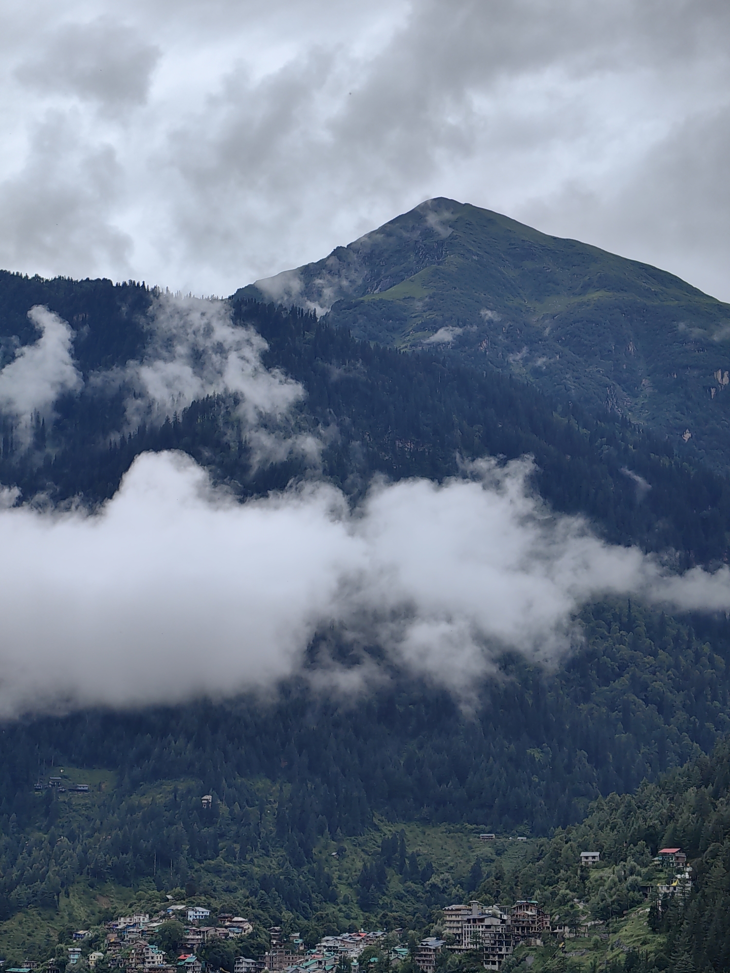 The image captures a dramatic mountain landscape under a cloudy sky, likely in or around Vashisht, India. The foreground features a cluster of buildings, representing a town or village nestled on a hillside. These structures are varied in size and architectural style, with some appearing to be multi-story hotels or residential buildings, while others are smaller houses. The hillside is densely covered with dark green coniferous trees, creating a textured backdrop for the settlement.
Mid-ground is dominated by a steep mountain slope, also heavily forested with the same dark trees. Wisps of white clouds drift and cling to the mountainside, obscuring parts of the dense foliage and adding a sense of depth and atmospheric conditions.
In the background, a larger, pointed mountain peak rises above the tree line. Its slopes are a mix of green, possibly covered with grass or low vegetation, and darker, rocky sections. Smaller pockets of clouds are also visible around this peak, some partially covering its summit. The overall sky is overcast with heavy, gray clouds, suggesting a potentially rainy or misty day. There are no visible people or specific actions depicted in the scene. The environment is characterized by its natural ruggedness, dense vegetation, and the presence of human settlement adapting to the mountainous terrain.