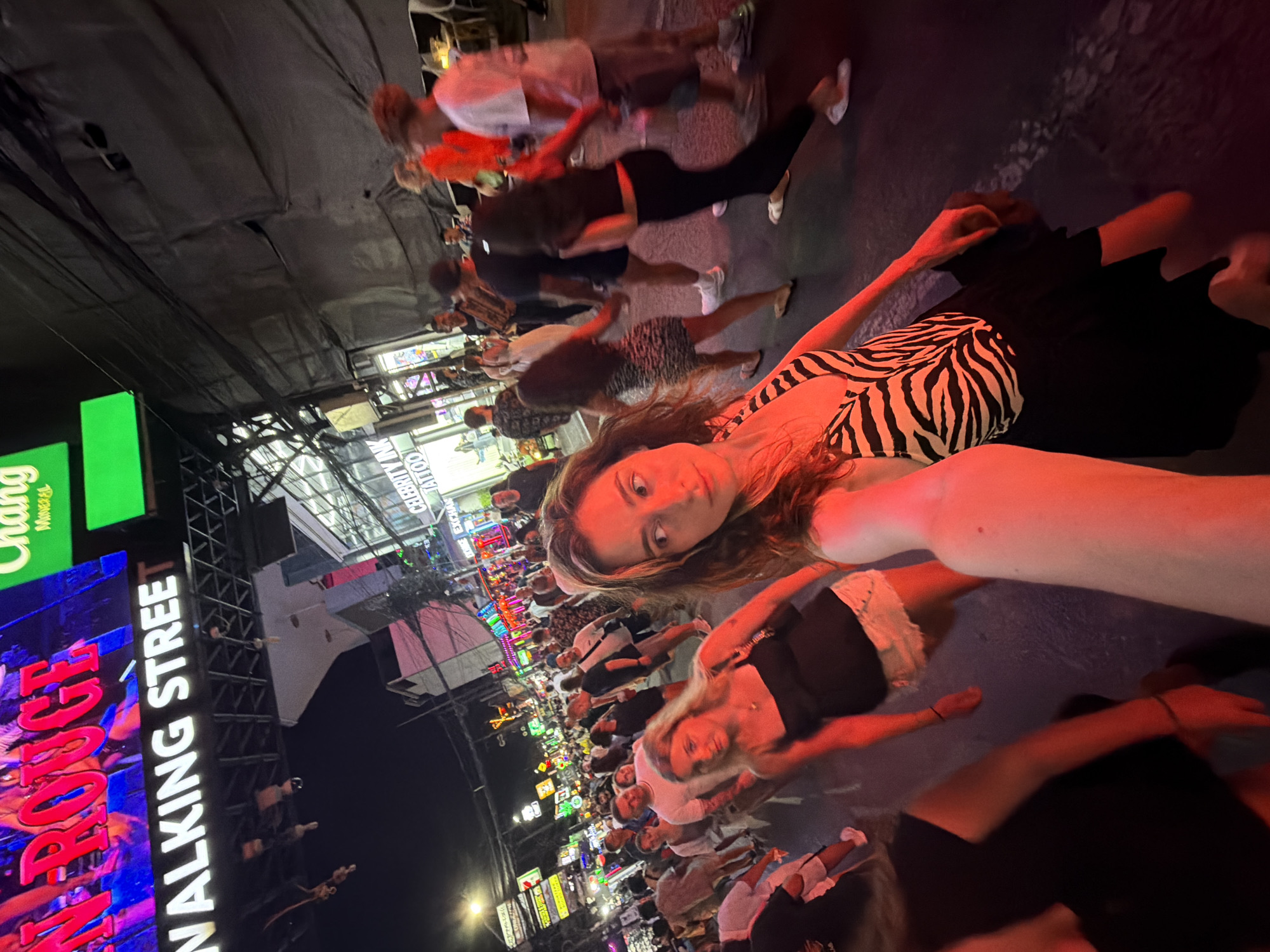 The image depicts a crowded nighttime street scene on Bangla Road in Patong, Phuket, Thailand, identifiable by the prominent "WALKING STREET" sign overhead and a "Chang Mineral" beverage sign. In the immediate foreground, a female individual with long brown hair, wearing a black and white zebra-print top and a black skirt, is capturing a low-angle selfie, illuminated by a strong red light source. Her face is partially visible, looking to her left. Slightly behind her and to her left, a blonde female in a black tank top and white shorts walks, also under red illumination. The remainder of the street is densely populated with a diverse group of people, primarily adults, walking in various directions. The background features numerous brightly lit neon signs from commercial establishments, including a prominent "CELEBRITY INK TATTOO" sign, creating a vibrant, illuminated urban atmosphere. Overhead, a complex network of wires and structural elements is visible against the dark night sky.