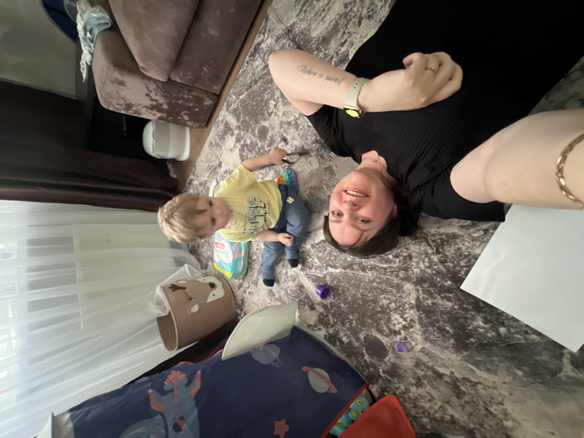 A woman and a young child are lying on a rug with a gray marble pattern. The woman is wearing a black dress and has a tattoo on her left forearm. She is taking a selfie. The child is wearing a yellow shirt and blue jeans. The rug is in a room with a brown couch, white curtains, and a toy bin with a deer pattern. There are various toys scattered around the rug. There is a box of Pampers diapers near the child. A blue play tent with planets is visible in the lower left corner. The location in the city of Noviy, Russia is not discernible from the image.