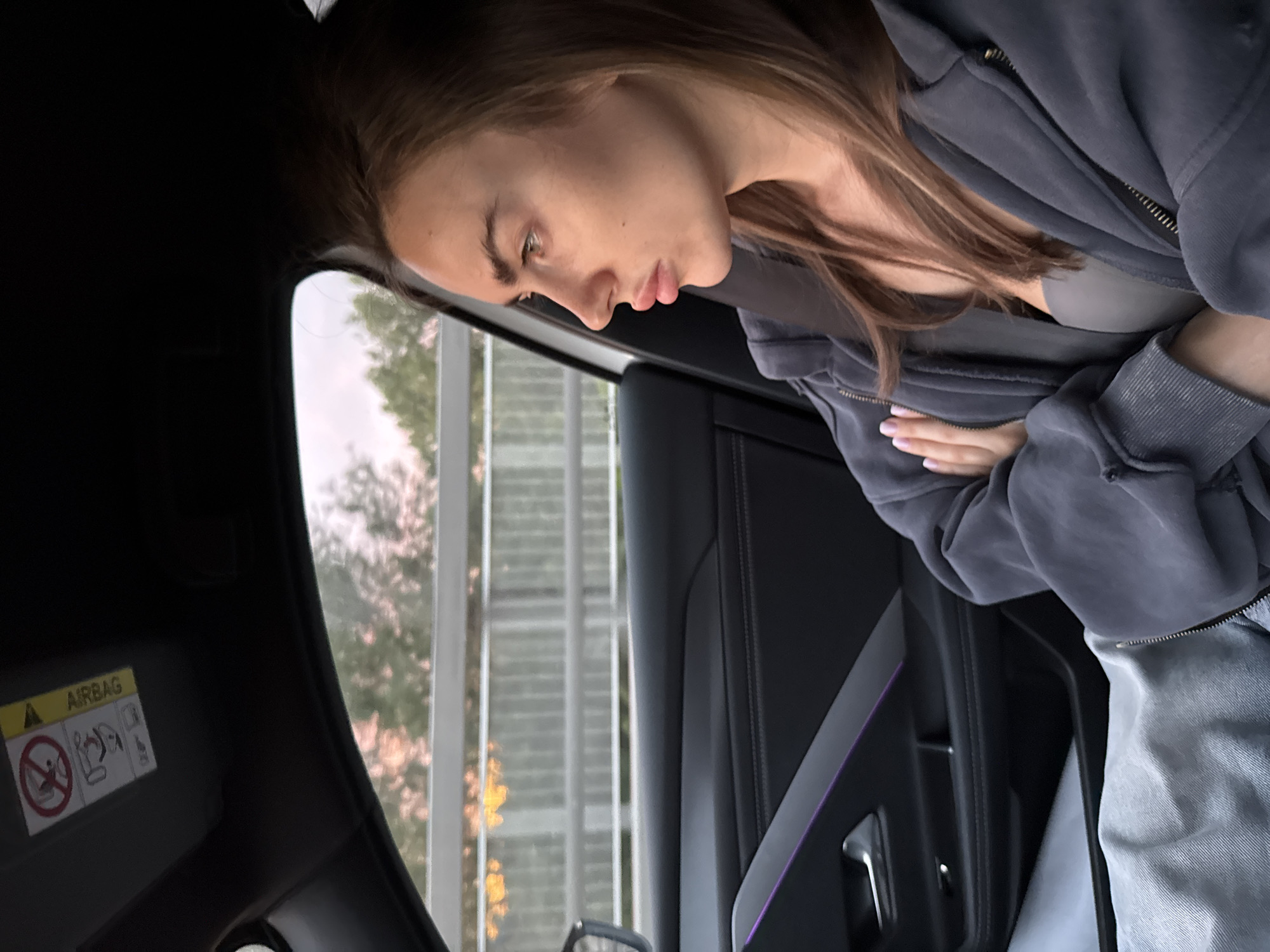 A young woman with long brown hair is sitting in the passenger seat of a car. She is wearing a gray zip-up hoodie and a seatbelt. Her arms are crossed. The car interior is black with a sliver of purple accent lighting on the door panel. Outside the window, a building with multiple floors can be seen, along with trees. An "Airbag" warning label is visible on the sun visor.
