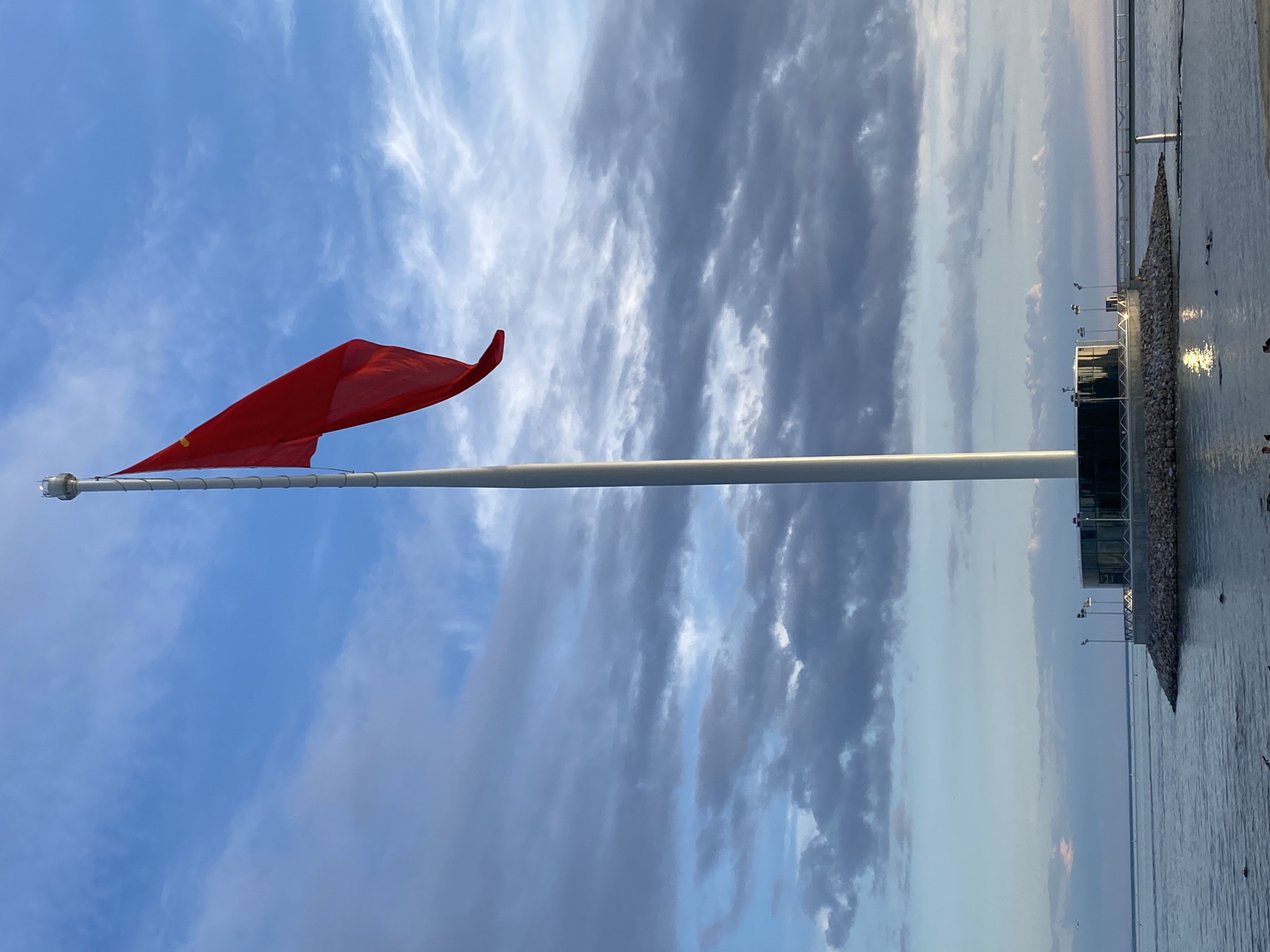 This image captures an expansive waterfront scene in Saint Petersburg, Russia, likely during early morning or late afternoon due to the soft, diffused lighting. A tall, white flagpole dominates the left side of the frame, bearing a plain red flag that billows to the right, indicating a gentle breeze.
The sky is a prominent feature, displaying a mix of bright blue on the left and scattered clouds ranging from wispy white to darker grey on the right. These cloud formations create a dynamic overhead canopy.
To the right, a calm body of water extends towards the horizon, reflecting the subdued light. Along the water's edge, a modern pier or embankment is visible. This structure features a stone or rocky shoreline leading up to several contemporary buildings with reflective surfaces. Multiple streetlights are also positioned along the pier. The overall impression is one of peaceful urban development blending with the natural expanse of the water and sky. No people or visible text are present in the image.