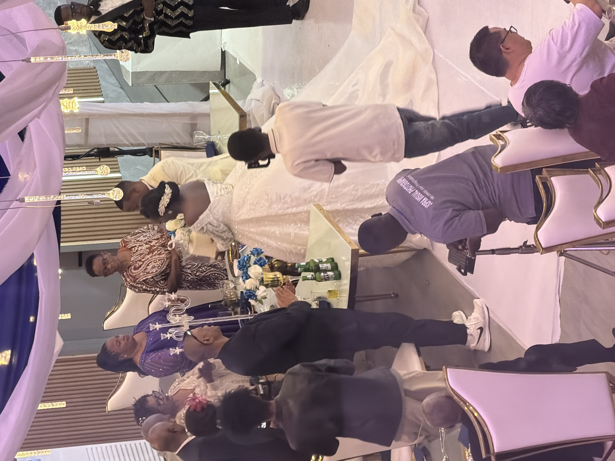This photograph captures a wedding reception in Dar es Salaam, Tanzania. The scene is set indoors, likely in a banquet hall, indicated by the draped fabric ceiling with decorative lighting, vertical paneling on the walls, and formal seating arrangements. Several individuals are present, dressed in celebratory attire.
In the foreground, a man in a dark suit and tie is standing near a table laden with items including bottles of beer, glasses, and a decorative centerpiece with flowers. To his right, a woman in a blue, heavily embellished dress is looking towards the center of the room. Behind her, another woman in a brown and gold patterned dress is also observing the proceedings. A bride, in a voluminous white wedding gown, is the central focus of attention in the mid-ground. She appears to be standing with a man in a cream-colored suit, possibly the groom.
Several other guests are visible throughout the image, some seated in ornate chairs, while others are standing and observing. A person in a gray t-shirt with visible text on the back is in the lower left quadrant, and another individual in a light pink t-shirt is in the lower right. The overall atmosphere suggests a formal event with a celebratory mood. The décor includes white tablecloths and chairs with gold accents, contributing to the elegant setting.