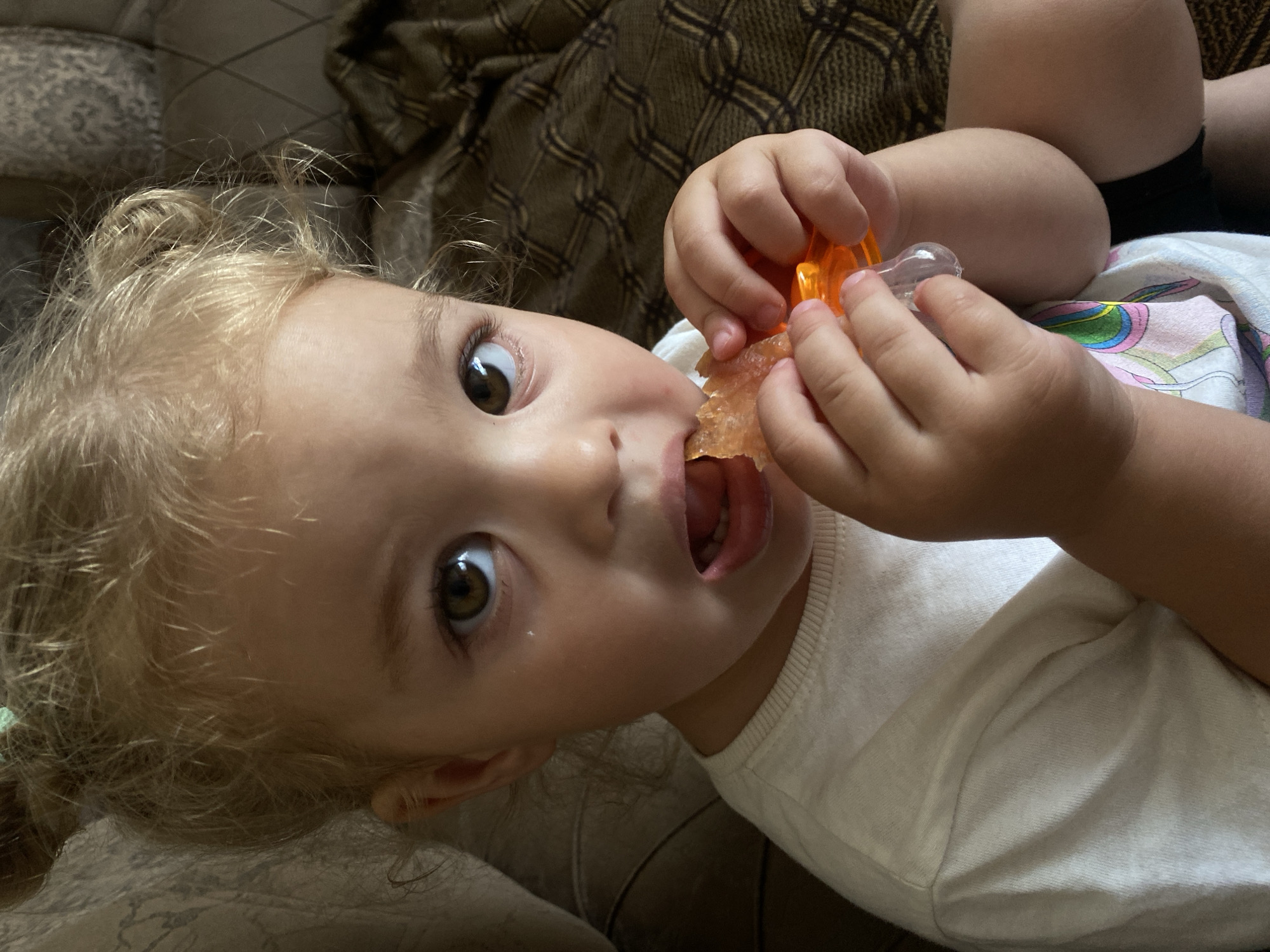 A close-up, eye-level shot shows a young child with blonde curly hair, a white t-shirt, and brown eyes. The child is sitting on a couch, holding an orange object with their hands and bringing it to their mouth. The child's mouth is open, and they appear to be eating or sucking on the object. The background is blurred, with hints of a patterned couch and blanket. The scene is indoors, likely in a home setting. The child's hair is styled in two messy buns.