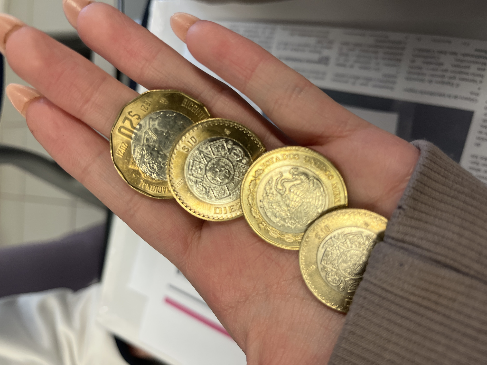A human hand, likely female, holds four Mexican bimetallic coins in a slight cascade. The hand exhibits light to medium skin tone and manicured, long, peach-colored fingernails. A light brown ribbed sleeve is partially visible on the lower left. The coins, from top to bottom (closest to fingertips to wrist), include: one $20 peso coin dated 2021, commemorating the Bicentennial of National Independence (1821-2021); one $10 peso coin displaying the Piedra del Sol (Aztec Sun Stone); one $10 peso coin showing the National Coat of Arms of Mexico with "ESTADOS UNIDOS MEXICANOS"; and a partially visible $10 peso coin also featuring the Piedra del Sol. The background is out of focus, revealing a blurred white surface on the left, and a white surface with indistinct text or markings on the right, suggesting an indoor setting. The scene is depicted as occurring in the city of Puebla, Mexico.