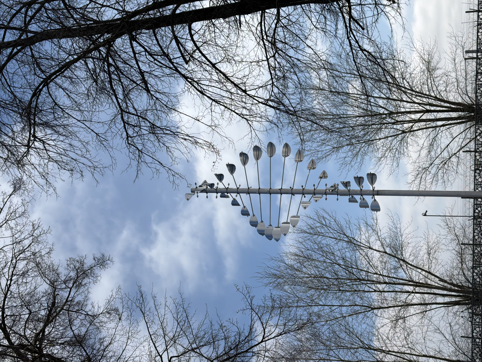 A low-angle shot depicts a kinetic sculpture, likely a wind chime or similar art installation, against a backdrop of a partly cloudy sky and bare tree branches. The sculpture consists of a horizontal pole from which a series of metal elements of varying shapes and sizes are suspended. These elements appear to be designed to move with the wind. The shot is framed by bare tree branches that spread out at the top and bottom of the image. The sky is visible through the branches and around the sculpture. The location is in the city of Votkinsk, Russia.